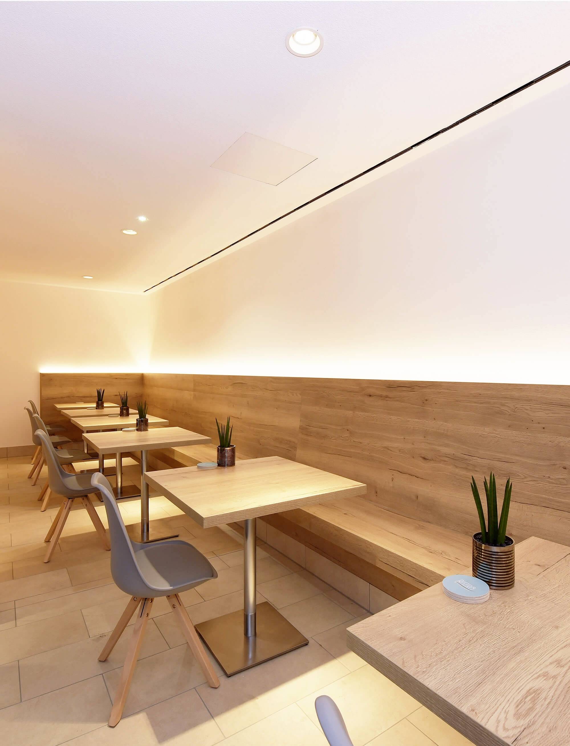 Un caffè moderno con tavoli in legno chiaro e sedie grigie; piccole piante decorano i tavoli, l'illuminazione a parete crea un'atmosfera accogliente.
