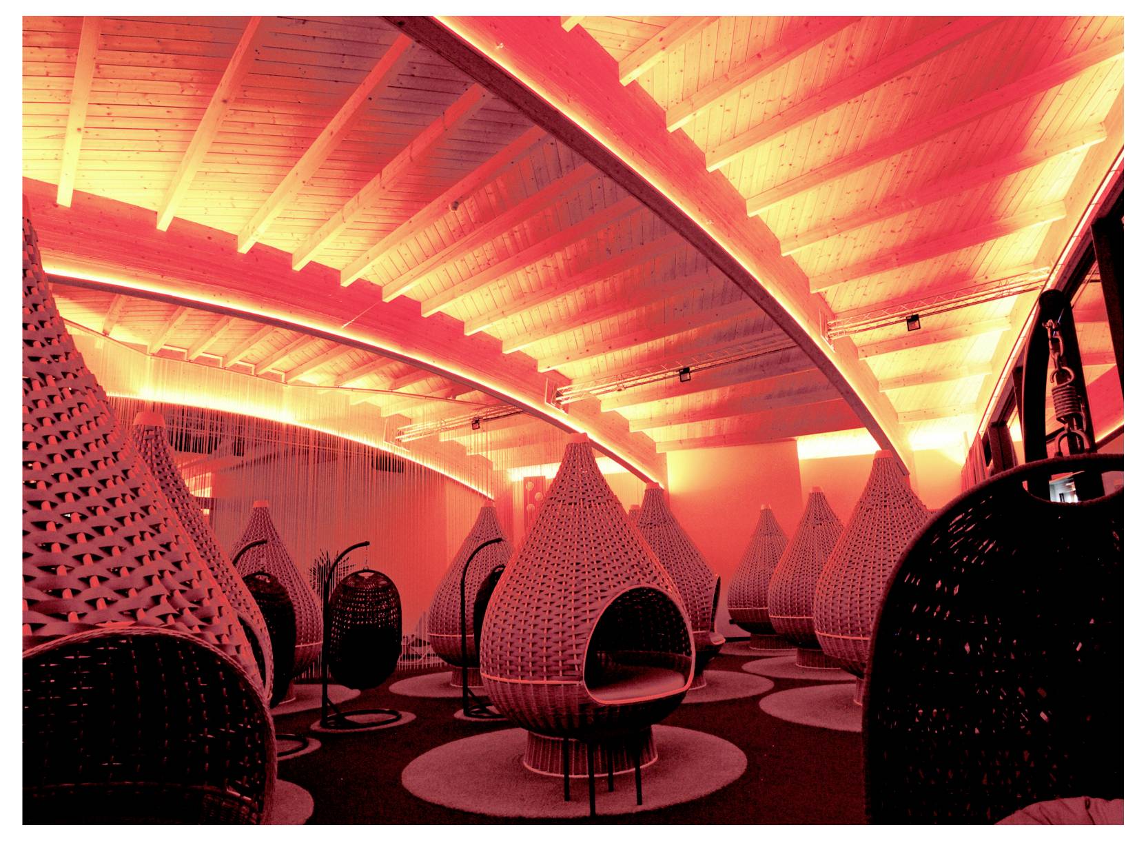 Salotto futuristico con sedie a sospensione intrecciate a forma di baccello e illuminazione curva a soffitto, in un interno illuminato di rosso-arancio.