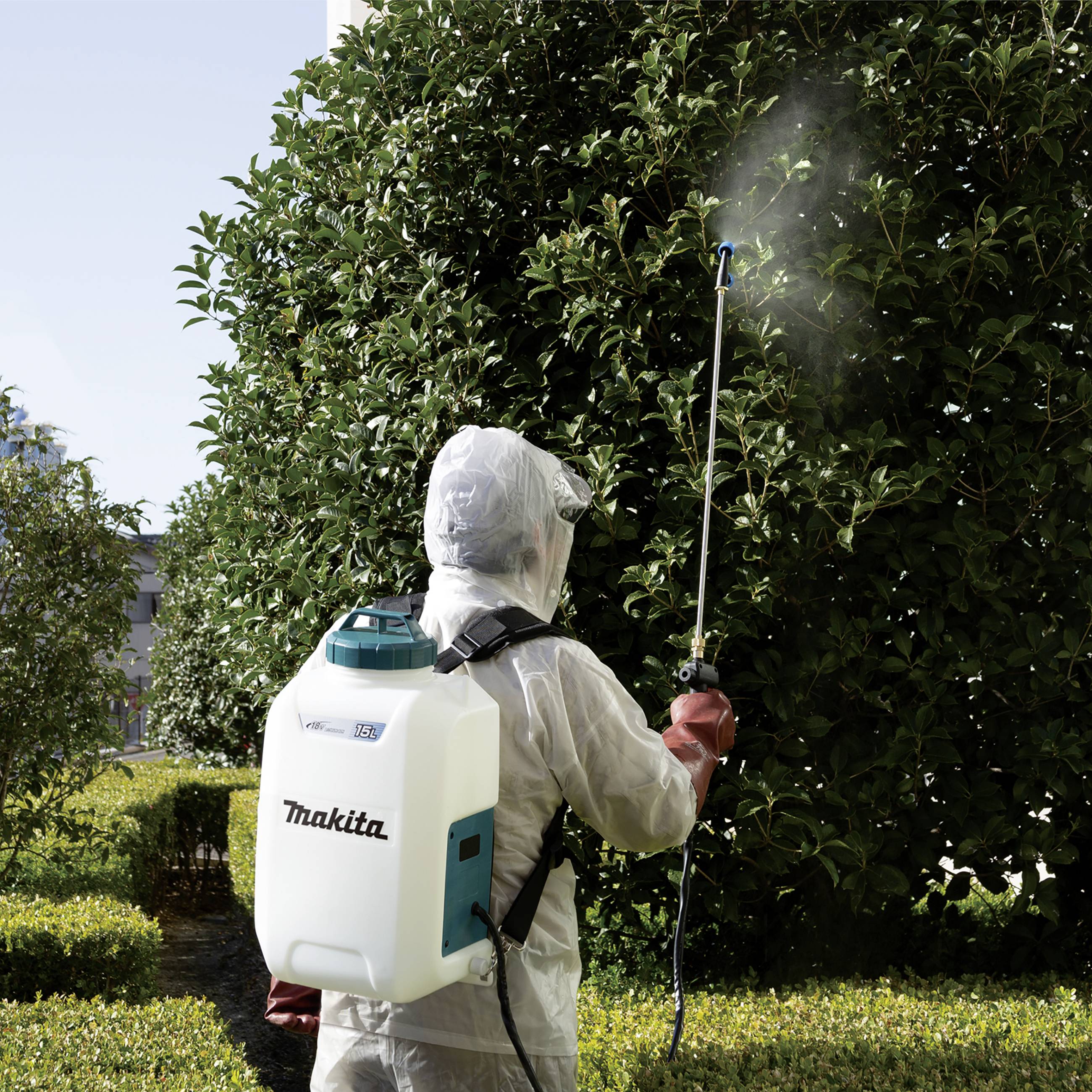 Una persona in tuta protettiva spruzza un cespuglio grande nel giardino con un irroratore a zaino. Il cielo è sereno.
