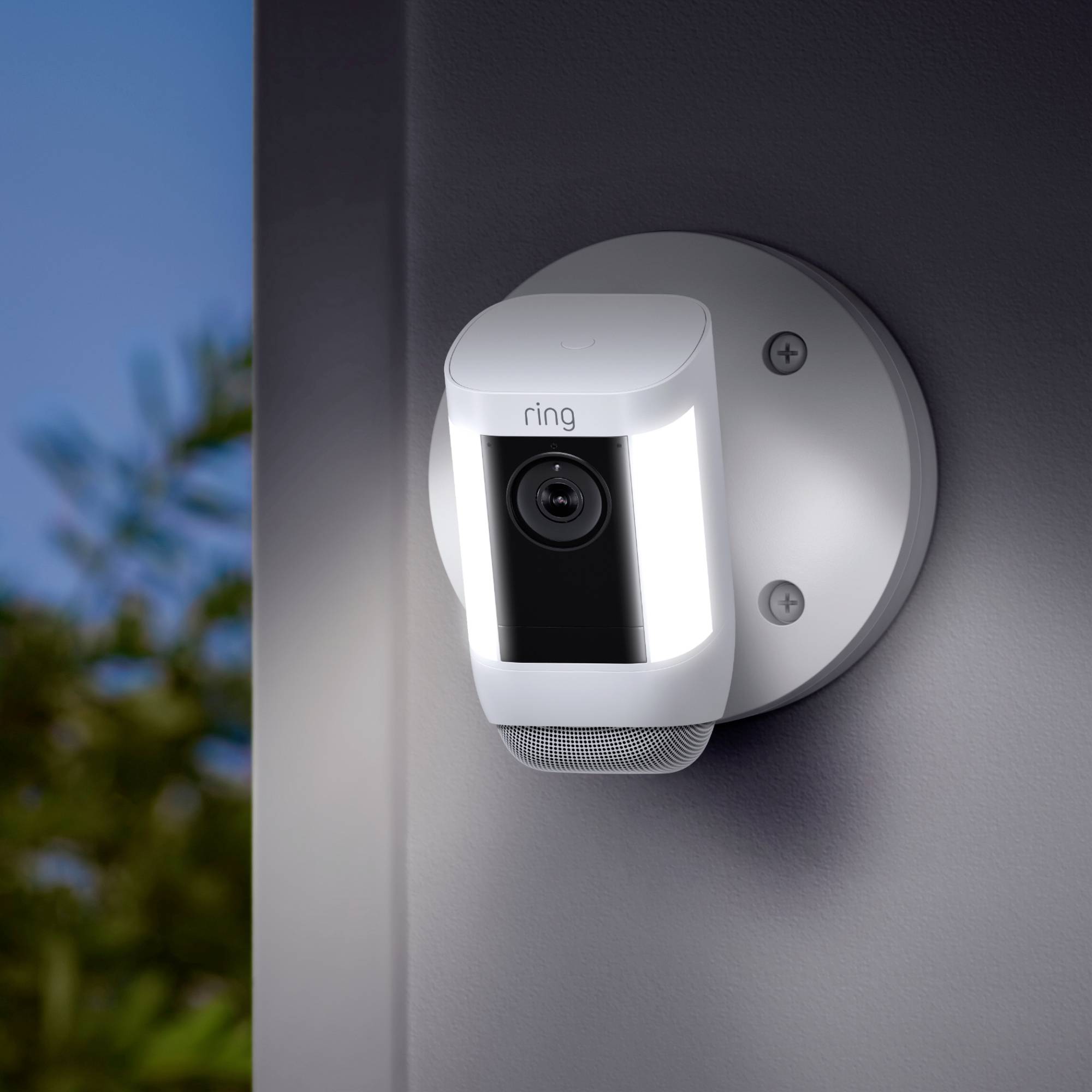 Telecamera di sorveglianza con luce LED e altoparlante montata su un muro, rivolta verso l'esterno in direzione degli alberi.