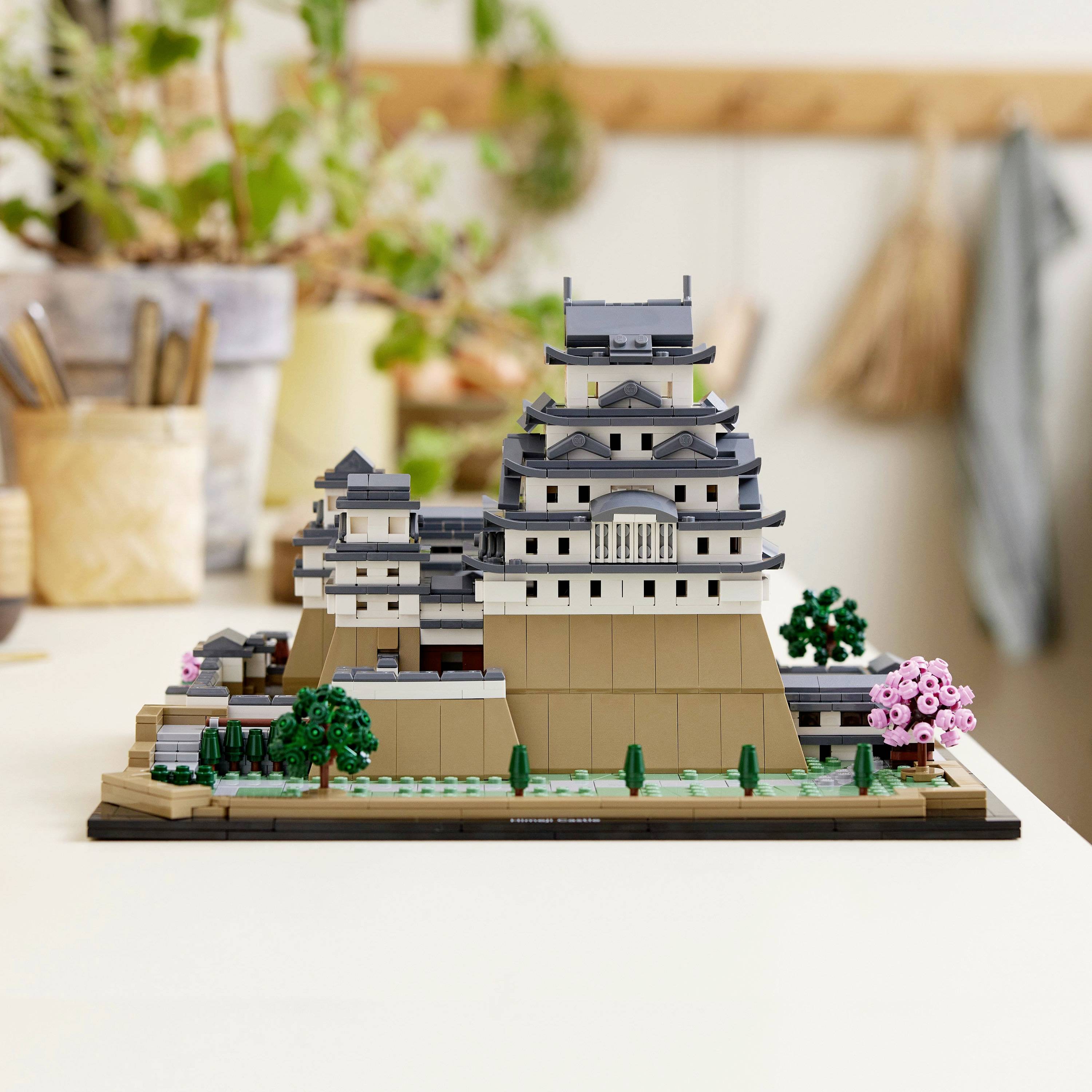 Modello Lego di un castello giapponese su un tavolo con piante sullo sfondo. Design ben strutturato con alberi.