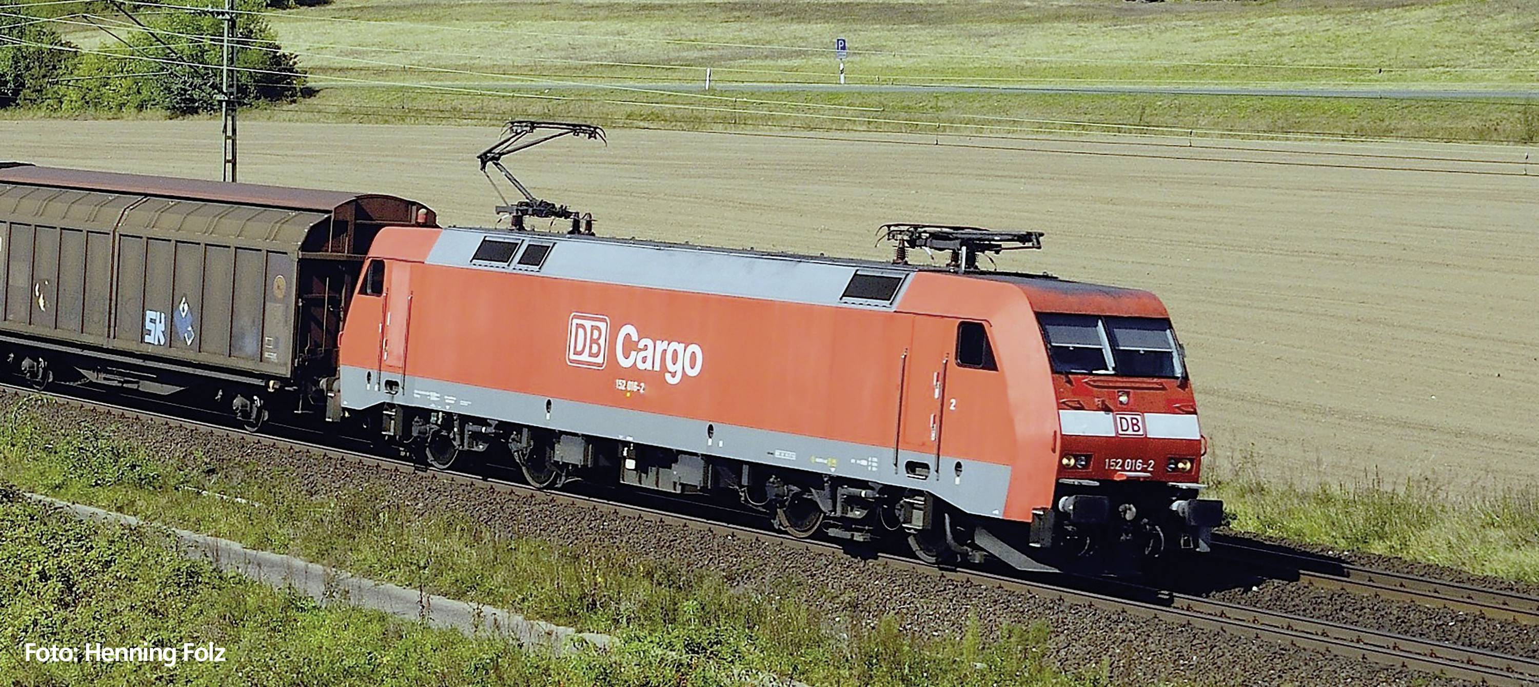 Un treno merci rosso di DB Cargo viaggia su binari attraverso un paesaggio rurale durante il giorno. A sinistra dell'immagine si trova un vagone merci.