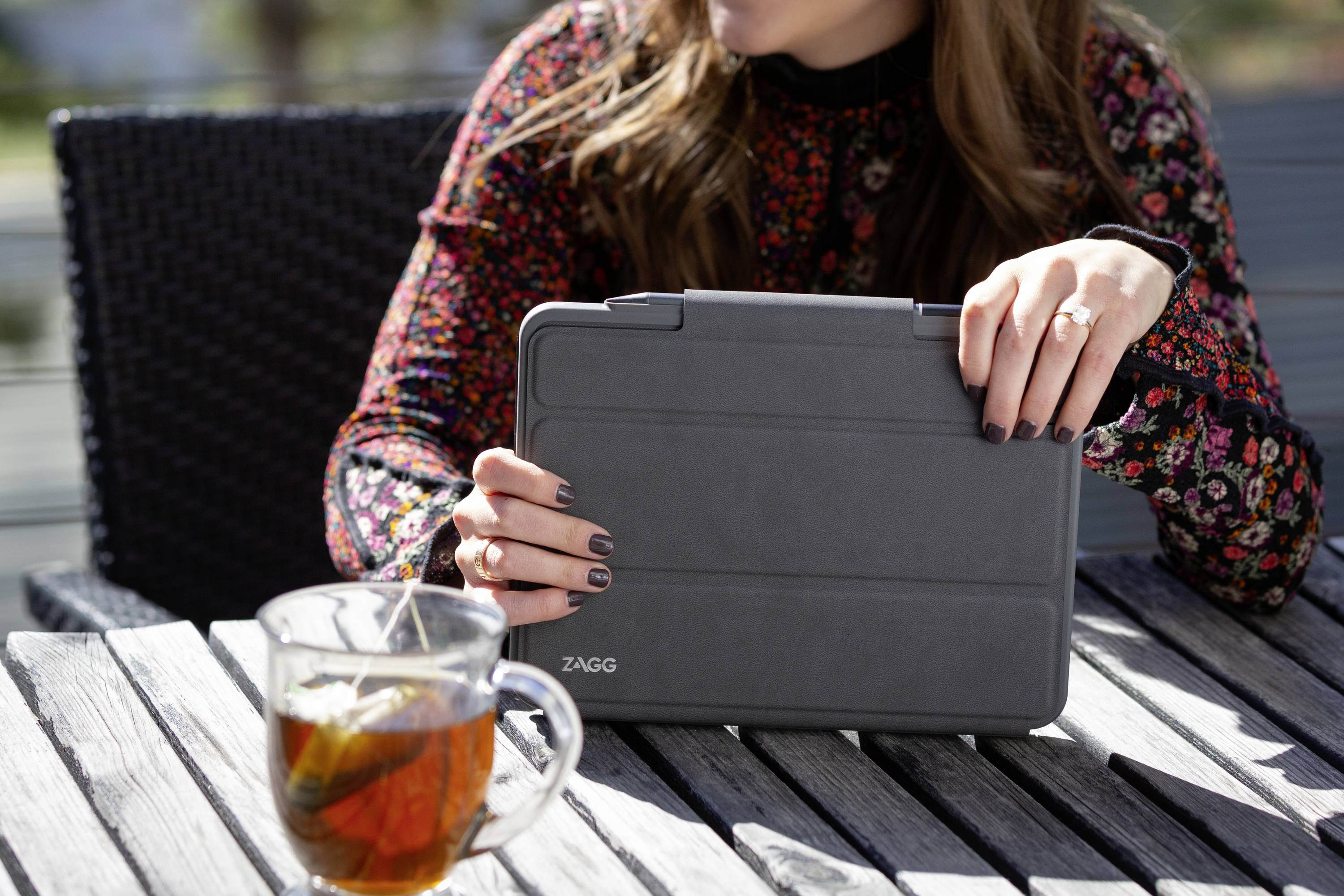 Una persona è seduta all'esterno a un tavolo e tiene in mano un tablet chiuso con una custodia nera. Sul tavolo c'è un bicchiere di tè.