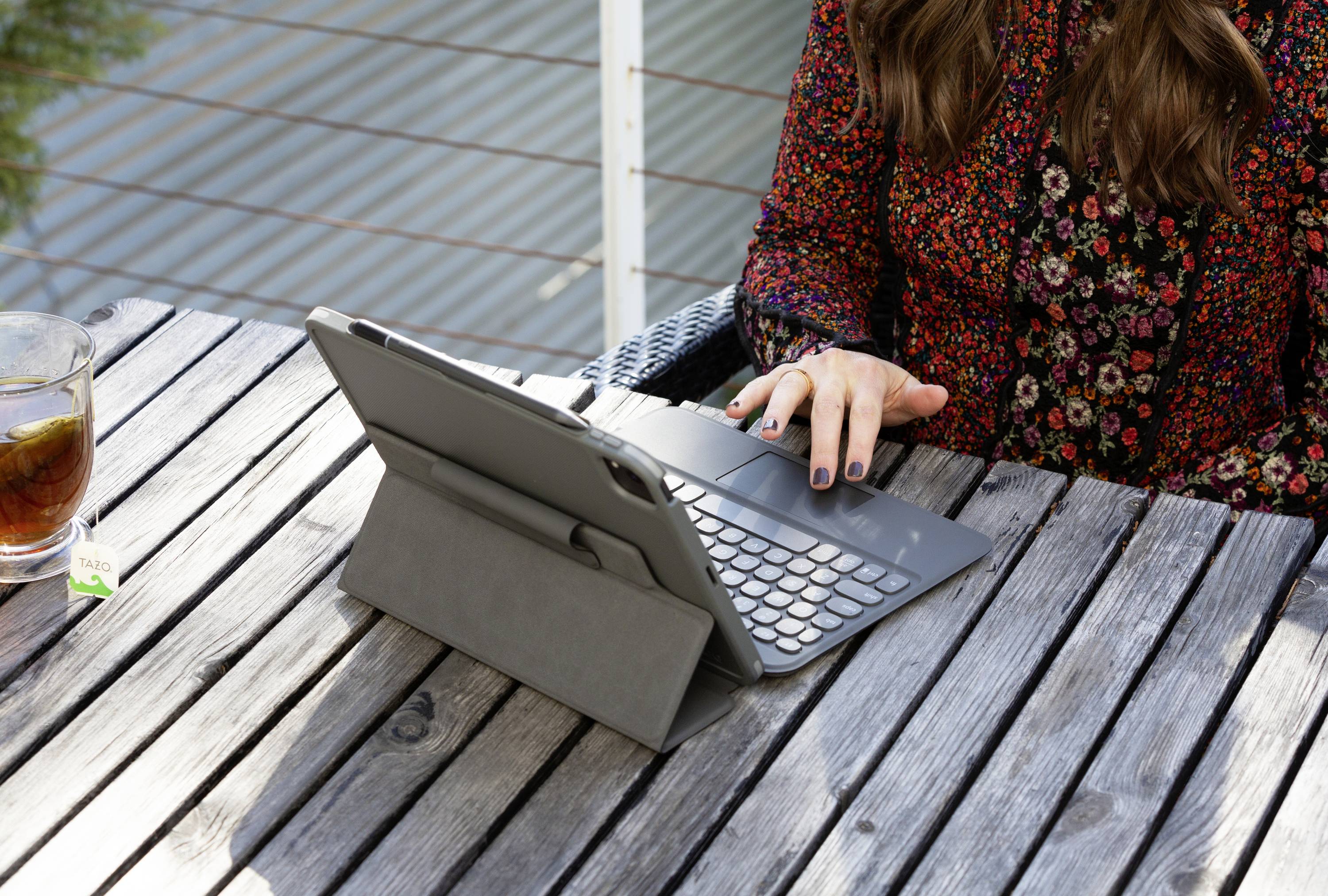 Una persona seduta a un tavolo di legno all'aperto con un tablet e una tastiera, accanto a un bicchiere di tè freddo.