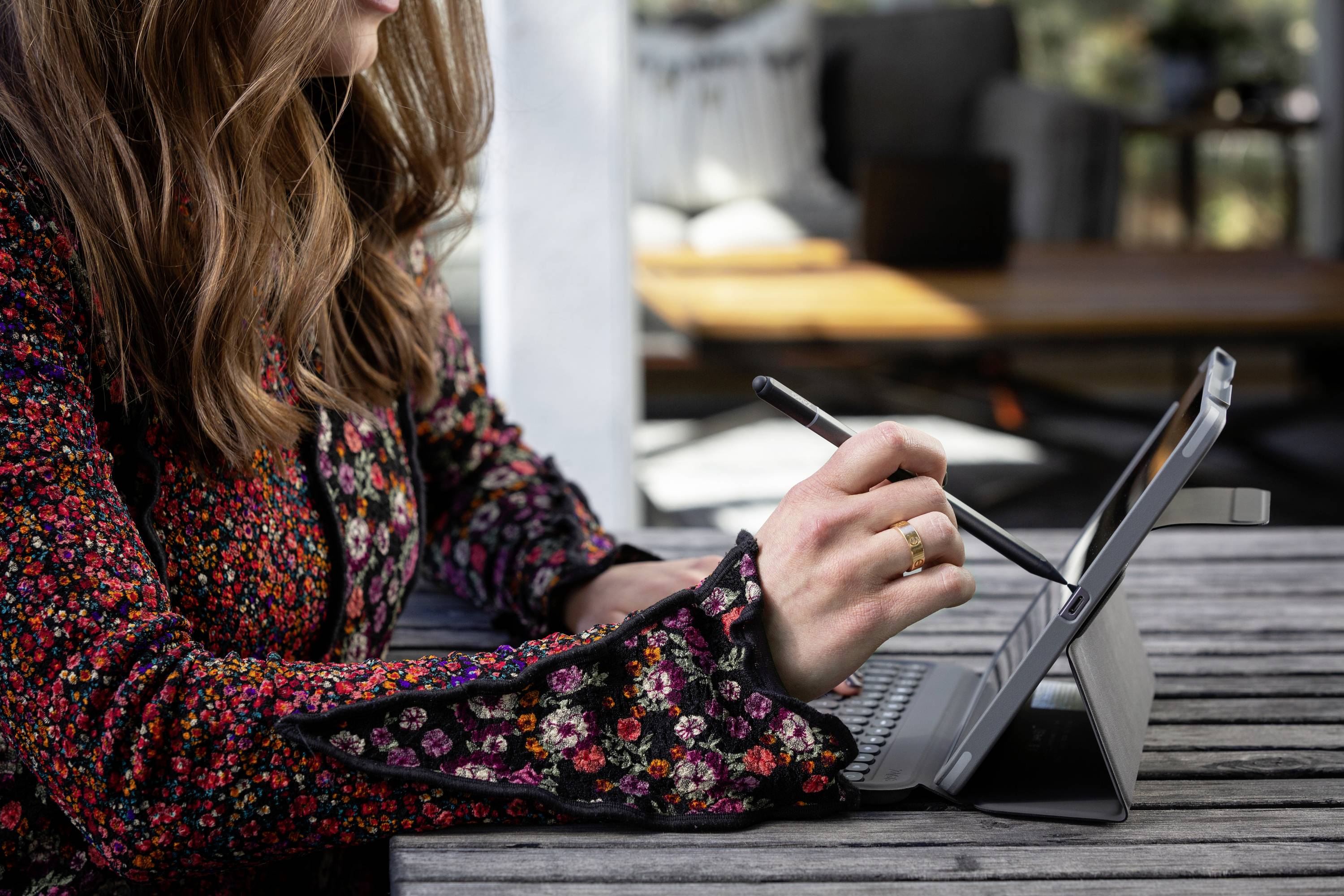 Una persona con un blazer colorato sta scrivendo con una penna su un tablet poggiato su un tavolo di legno. Sullo sfondo, mobili sfocati.