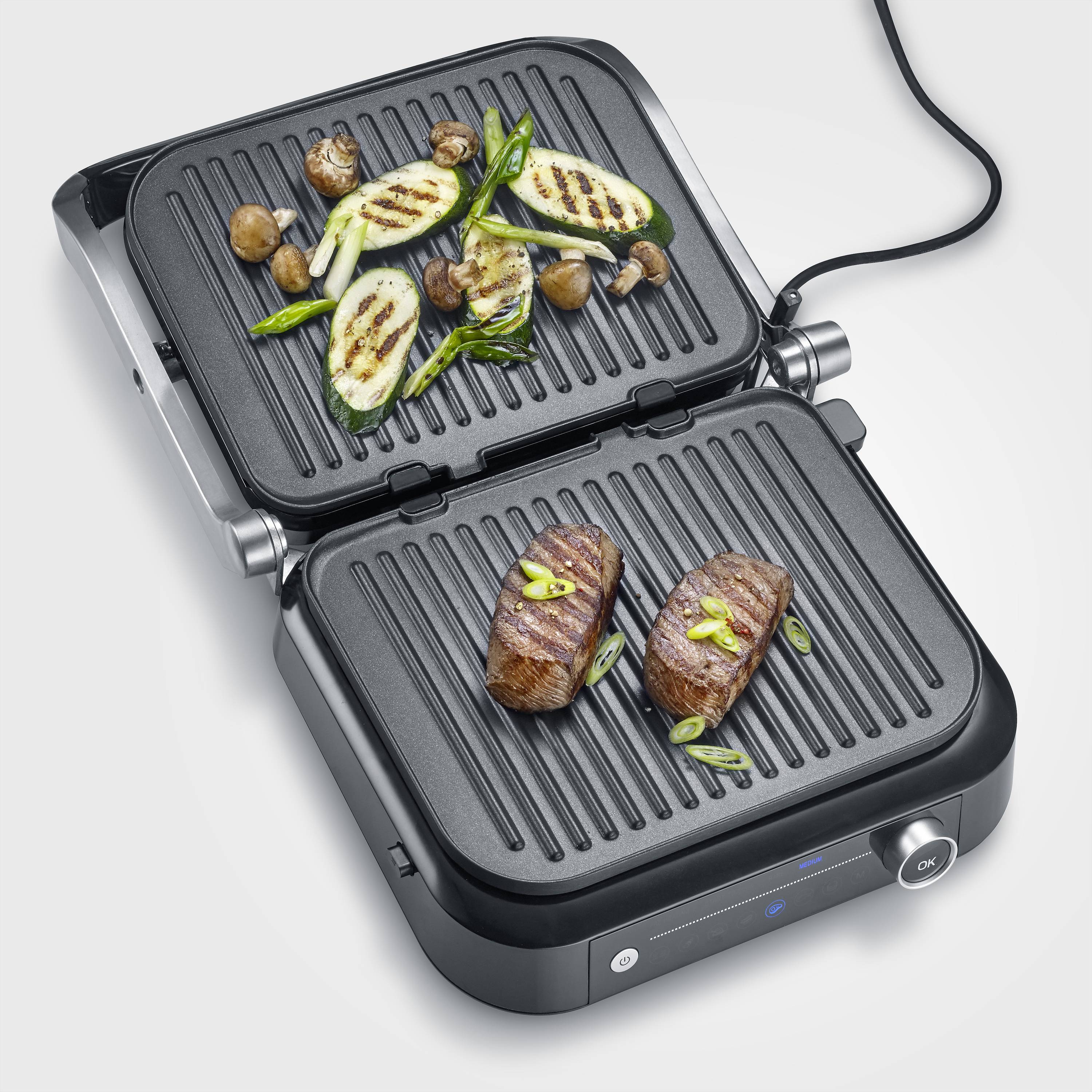 Un grill a contatto con bistecca alla griglia, zucchine, funghi e cipollotti. La griglia è elettrica e rappresentata aperta.