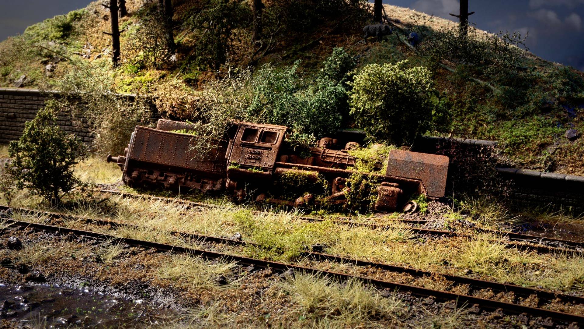 Una locomotiva arrugginita e capovolta giace accanto ai binari ferroviari in un ambiente rurale ricoperto di vegetazione.