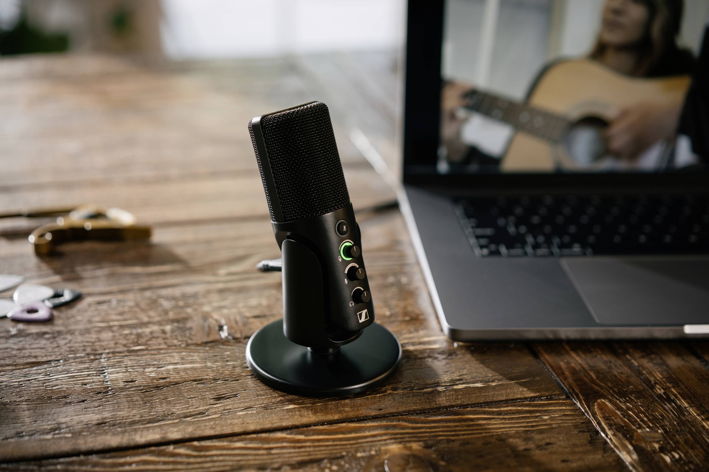 Un microfono è posizionato su un tavolo di legno accanto a un laptop, il cui schermo mostra una persona che suona la chitarra.