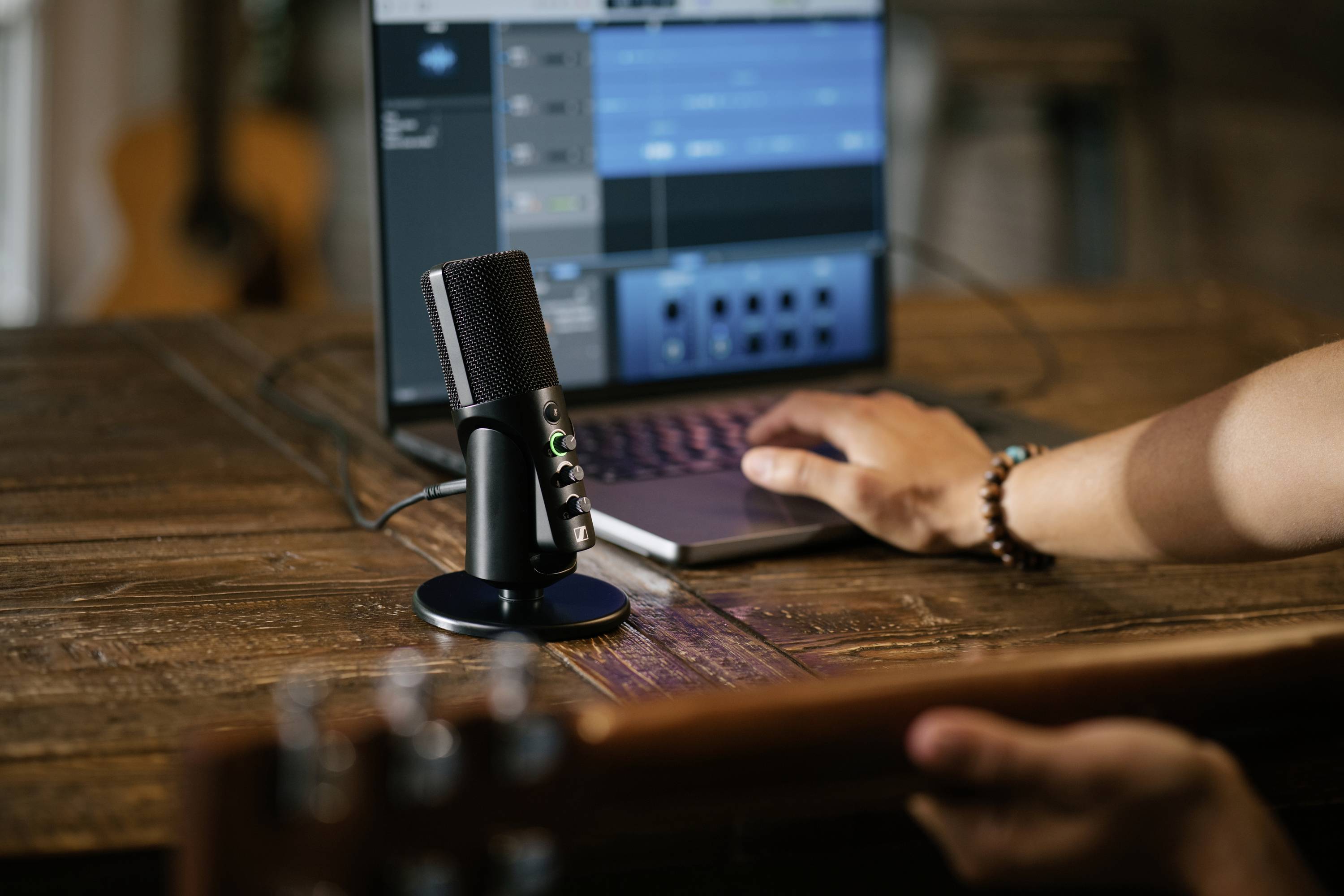 Un microfono su un tavolo davanti a un laptop mostra un programma audio. Una persona sta modificando file audio e suonando la chitarra.