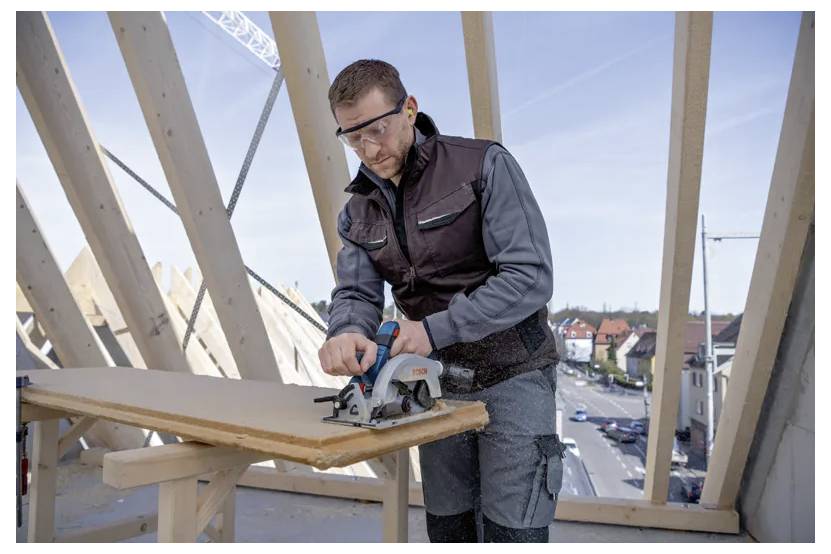 Una persona che indossa dispositivi di protezione aziona una sega circolare su un pezzo di legno in un cantiere edile, con una città visibile sullo sfondo.