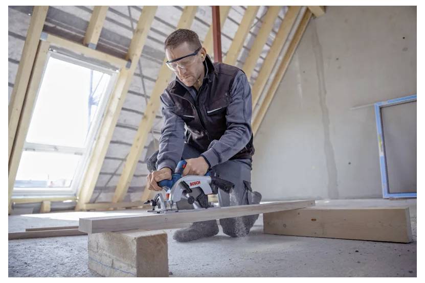 Una persona che indossa occhiali di protezione utilizza una sega elettrica per tagliare un'asse di legno in uno spazio sottotetto non finito, con la luce che entra da una finestra.