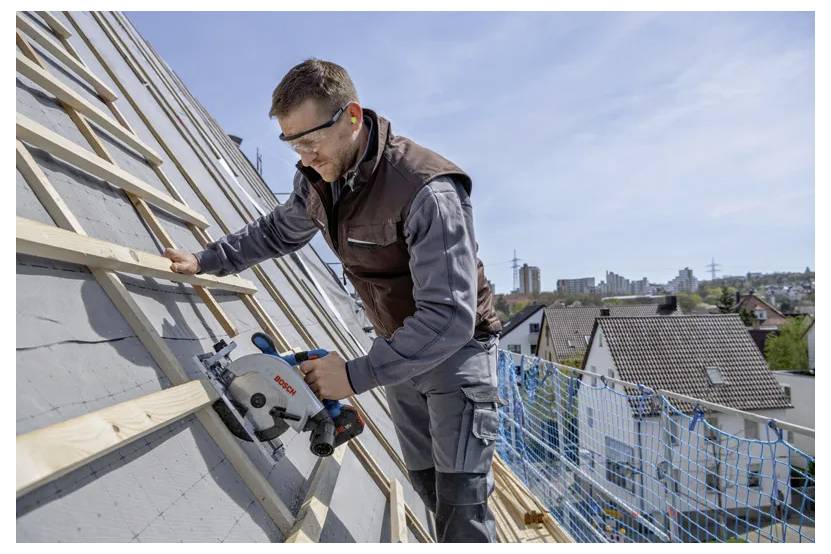 Lavoratore su tetto inclinato che utilizza una sega circolare per tagliare un listello di legno, indossa occhiali di protezione e giubbotto di sicurezza, con edifici cittadini sullo sfondo.