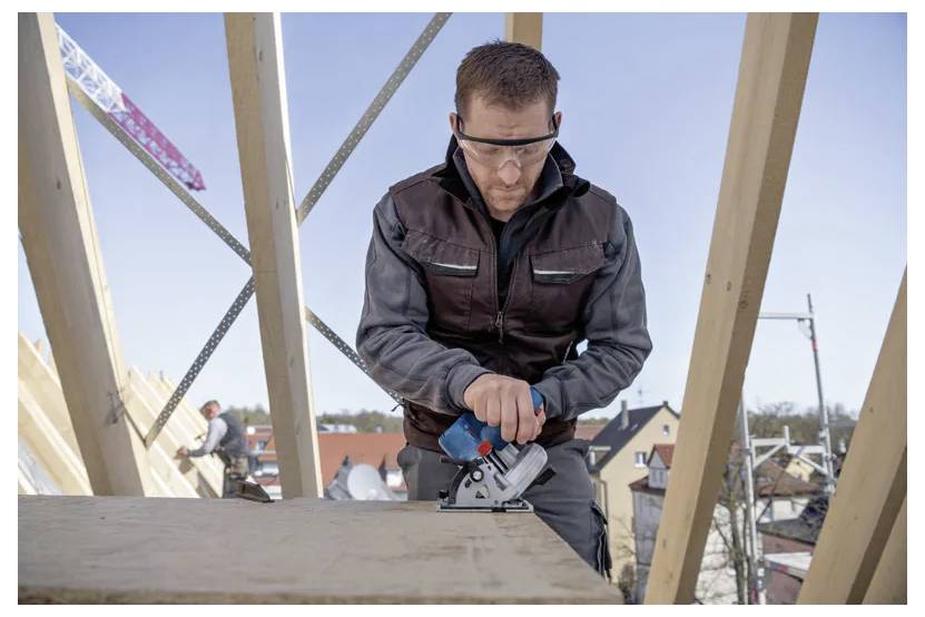 Un lavoratore edile che indossa occhiali protettivi aziona un utensile elettrico su una trave di legno in un cantiere edile, con i tetti visibili in basso.