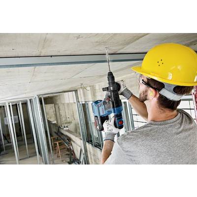 Un operaio edile con un casco giallo sta usando un trapano blu per lavorare su un soffitto. Sullo sfondo si vede un'impalcatura metallica.
