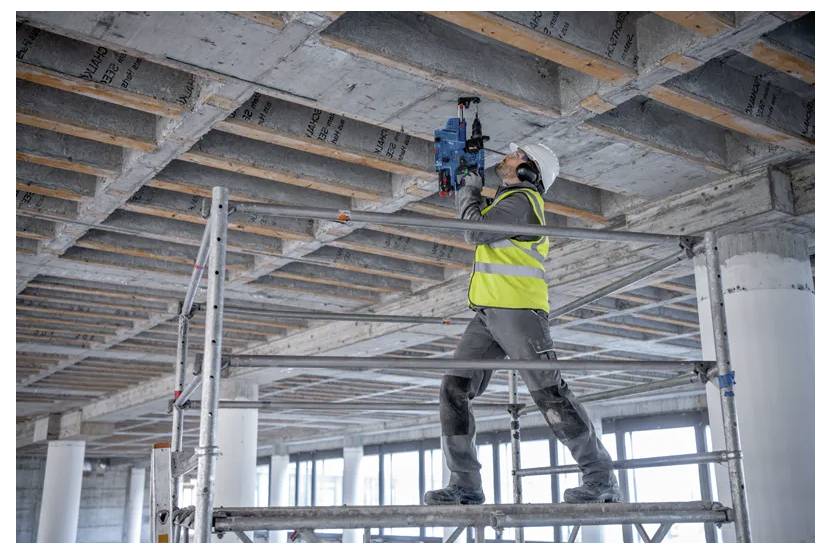 Un lavoratore edile con un giubbotto ad alta visibilità e un elmetto è in piedi su un ponteggio, utilizzando un utensile elettrico su un soffitto in calcestruzzo all'interno di un edificio.