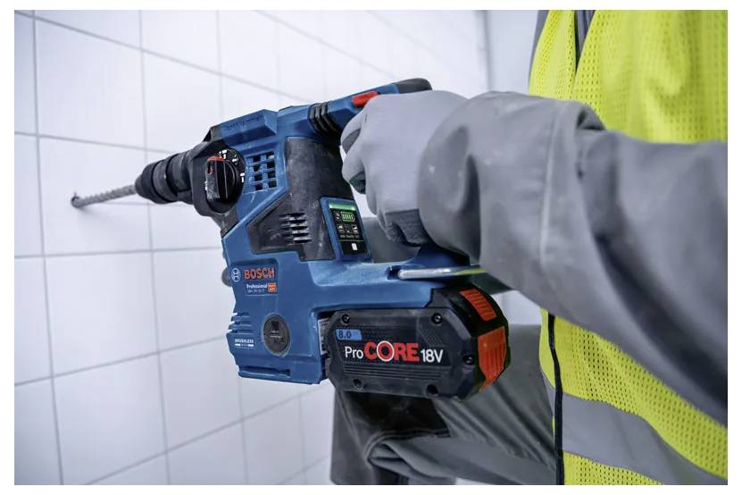 Una persona che indossa un giubbotto di sicurezza giallo utilizza un trapano Bosch ProCORE18V blu per forare una parete bianca piastrellata.