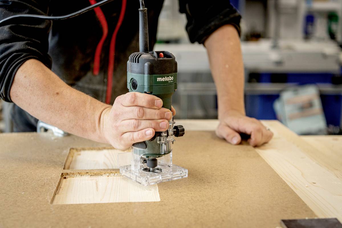 Una persona sta fresando un foro rettangolare in una tavola di legno utilizzando una fresatrice in un laboratorio.