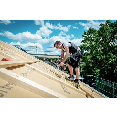 Un operaio edile con abbigliamento da lavoro è in piedi su un tetto inclinato, utilizzando un chiodatrice. Il cielo è nuvoloso, con alberi sullo sfondo.