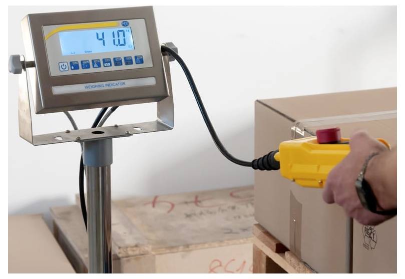 Indicatore di pesatura digitale che mostra 4.10 collegato tramite cavo a un dispositivo di controllo giallo accanto a un pacco inscatolato su un pallet.