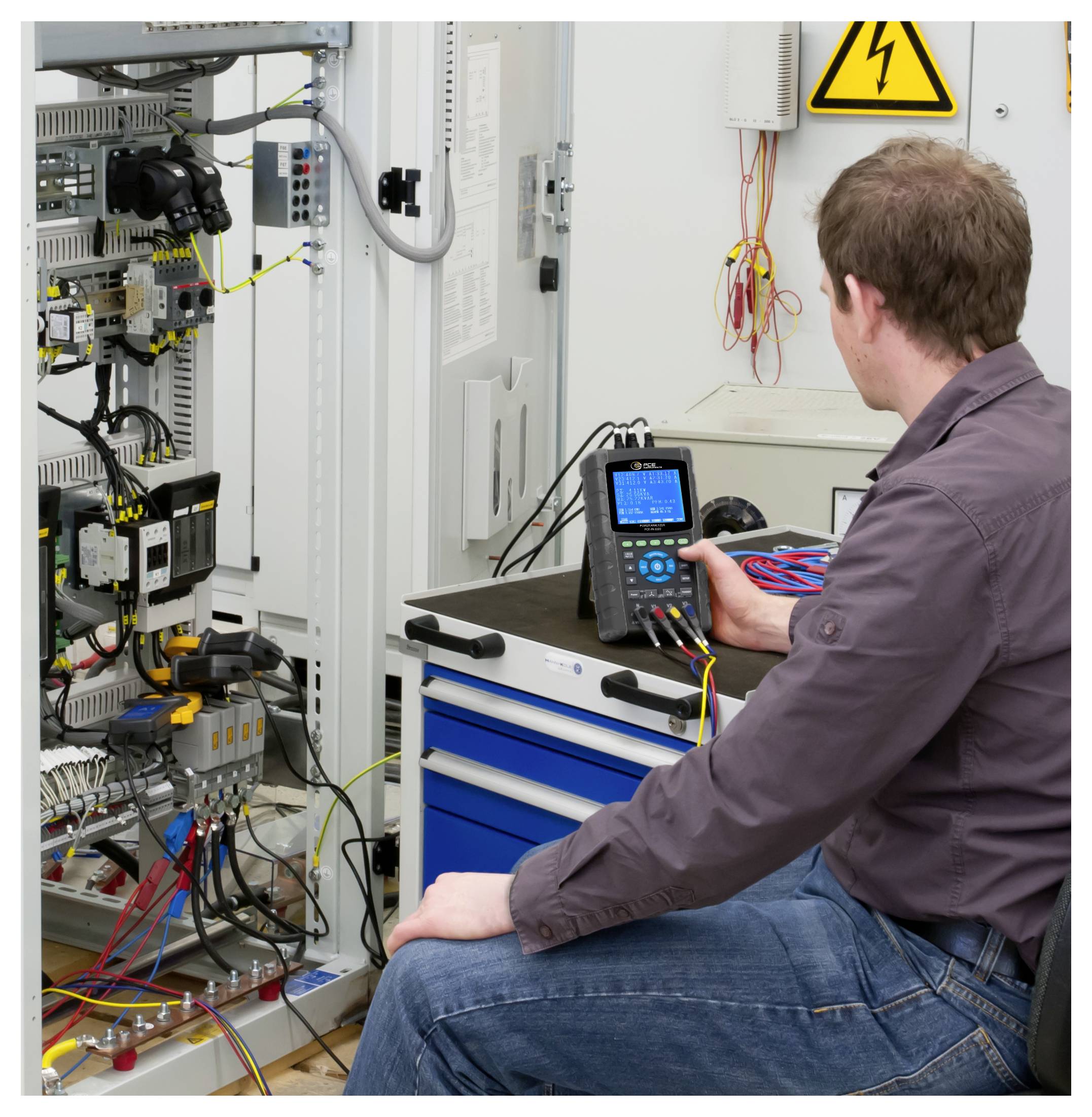 Un tecnico verifica il cablaggio di un quadro elettrico di comando con un multimetro portatile in un'officina, vicino a un cartello di avvertimento di alta tensione.