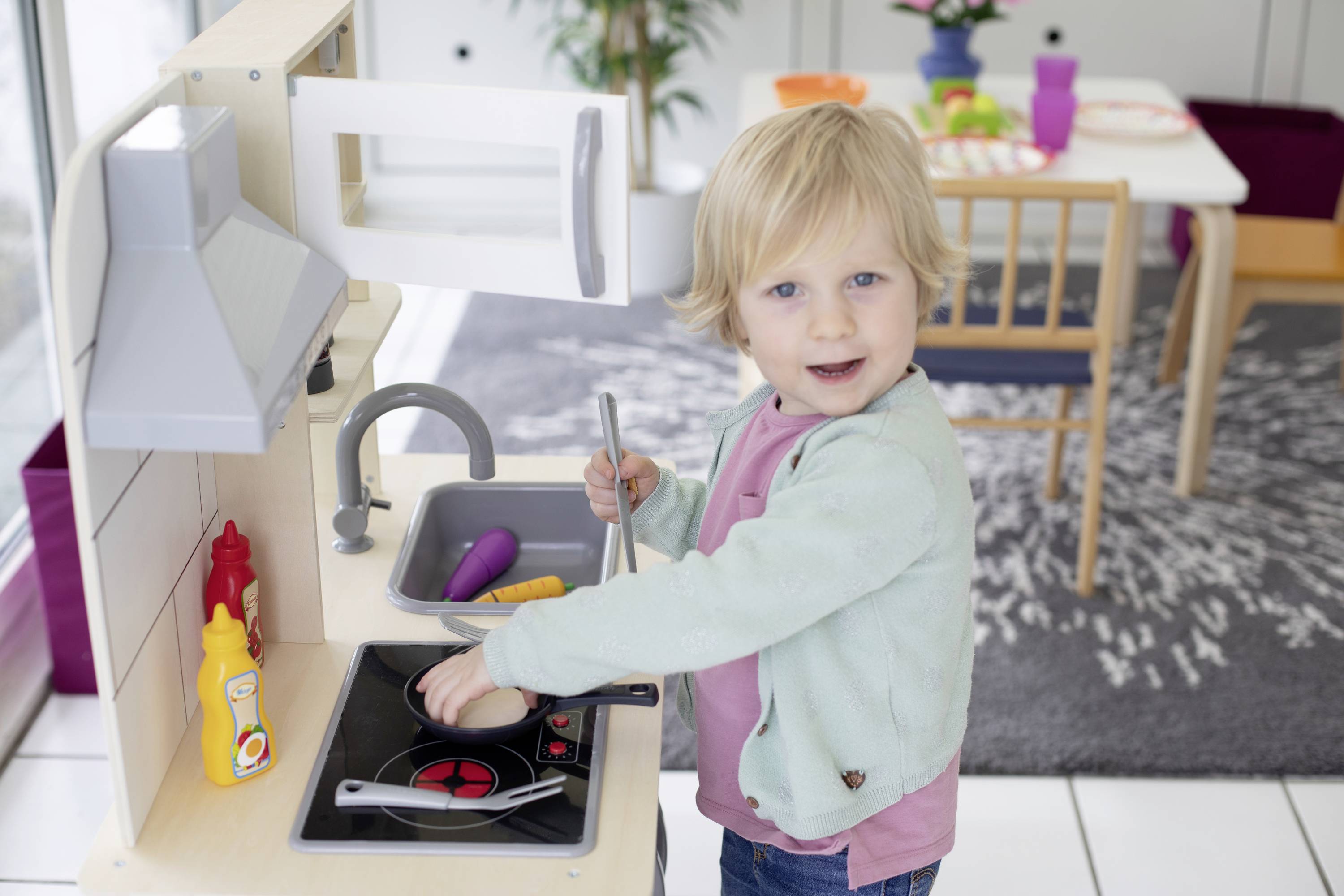 Un bambino gioca in una cucina giocattolo e usa una padella sul fornello. Sullo sfondo si vede un tavolo con stoviglie.