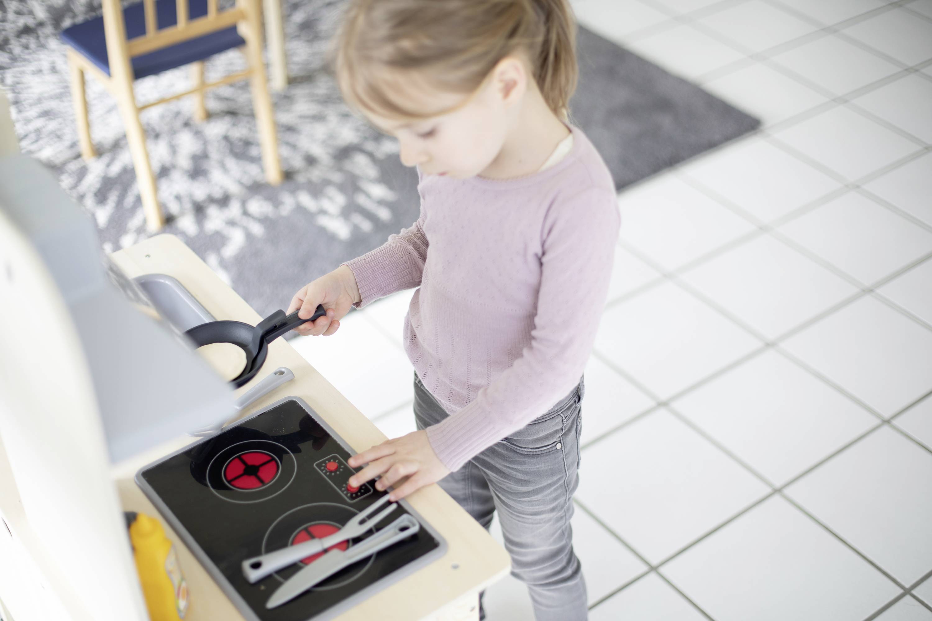 Un bambino gioca con una cucina giocattolo, cucina su una piastra da gioco con una padella in miniatura e delle posate, in un soggiorno.