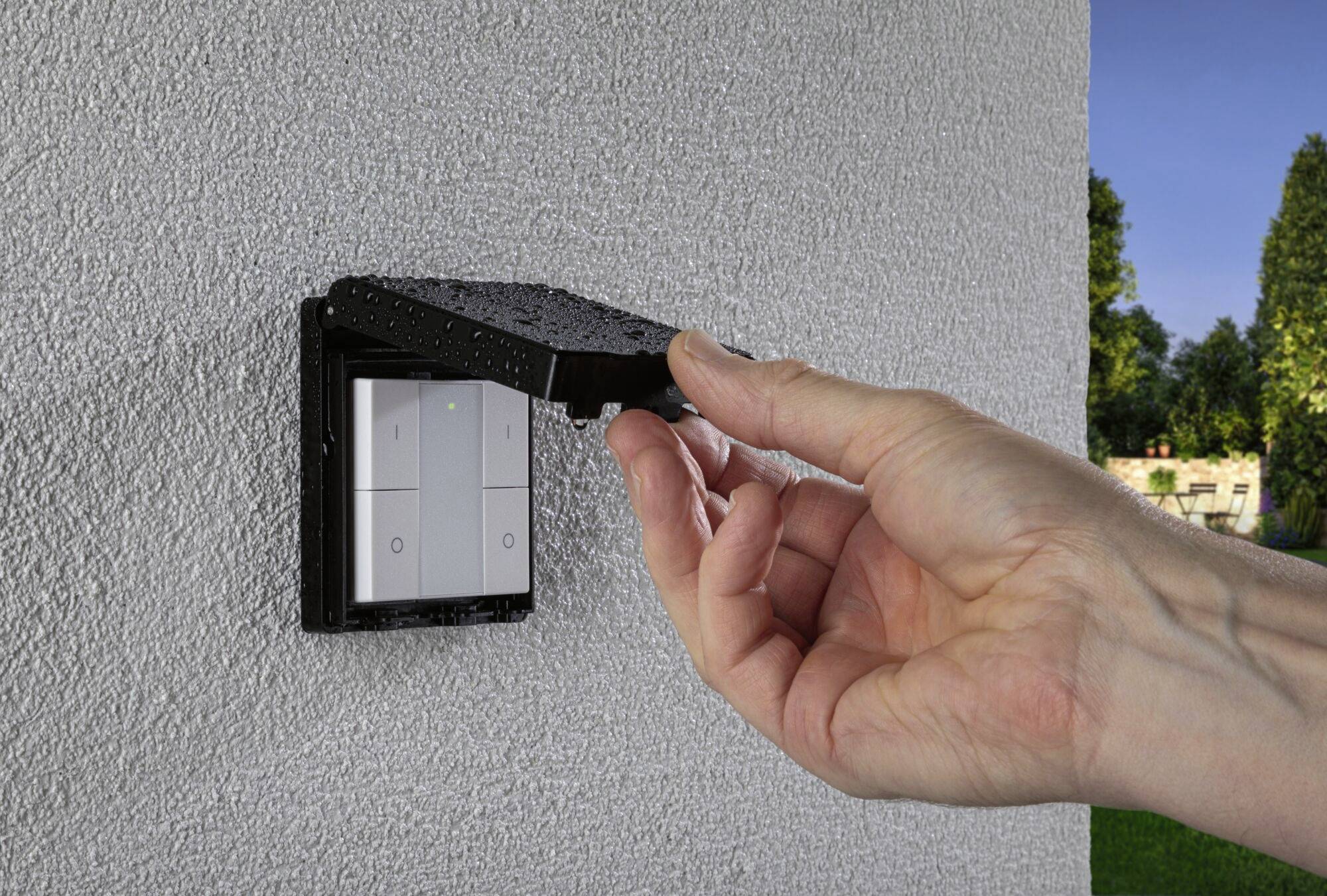 Una mano apre il coperchio di un interruttore elettrico su una parete esterna. L'interruttore è protetto dagli agenti atmosferici.