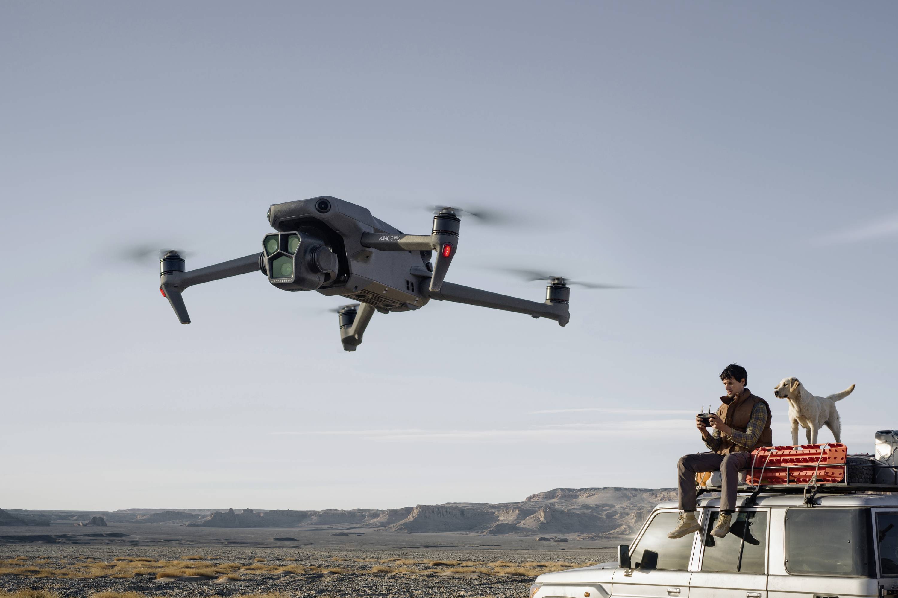 Un uomo è seduto su un fuoristrada all'aperto e pilota un drone, mentre un cane è in piedi accanto a lui. Paesaggio desertico sullo sfondo.