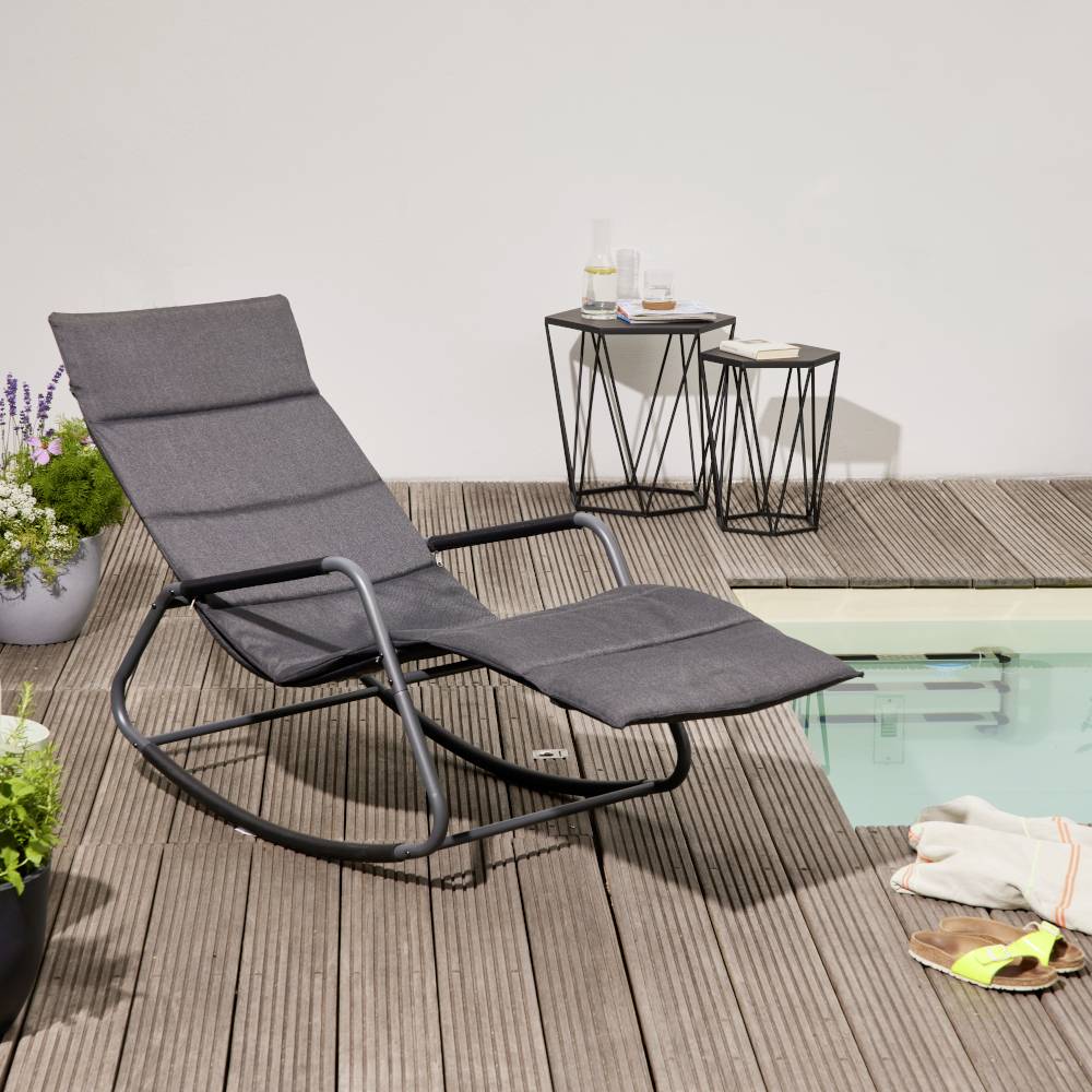 Una sedia a dondolo grigia è posizionata su una terrazza in legno accanto a un piccolo tavolo con bevande. Sullo sfondo, una piscina e alcune piante.
