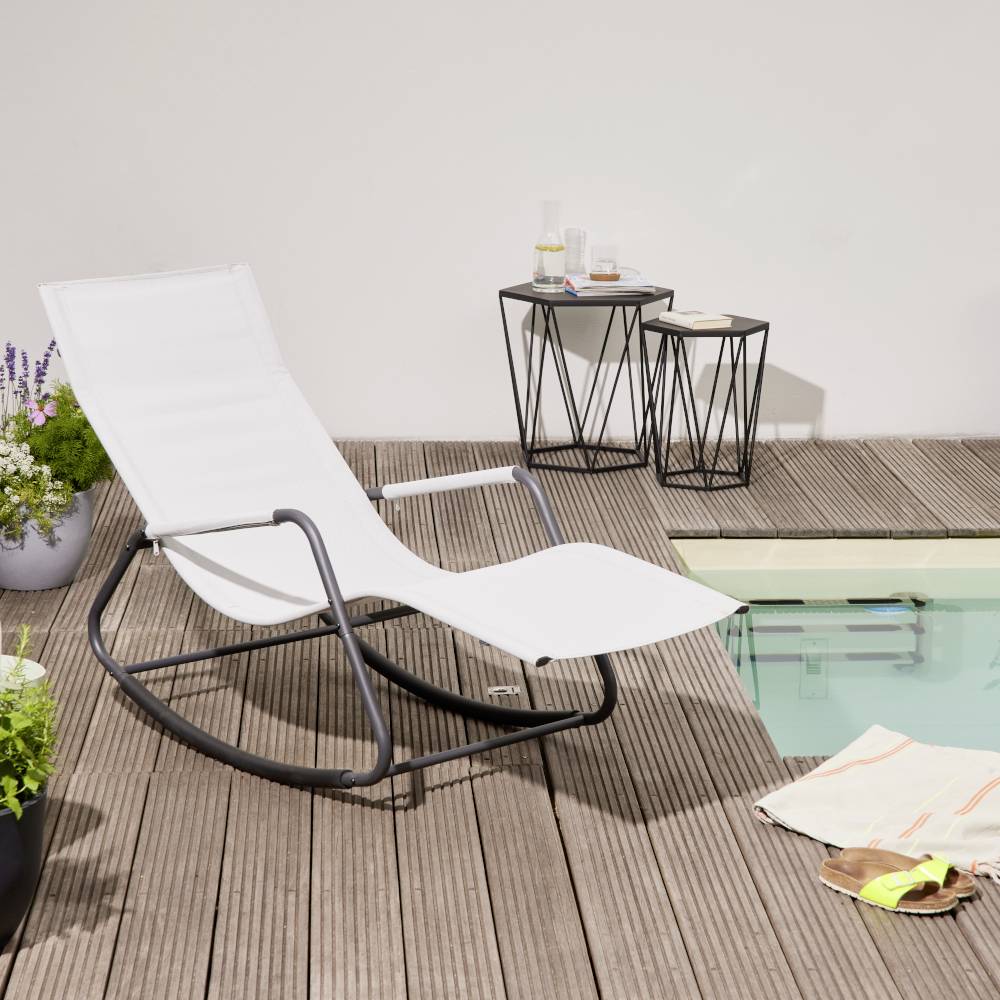 Lettino prendisole bianco e tavolino con bevande su una terrazza in legno accanto a una piccola piscina. Piante sullo sfondo.