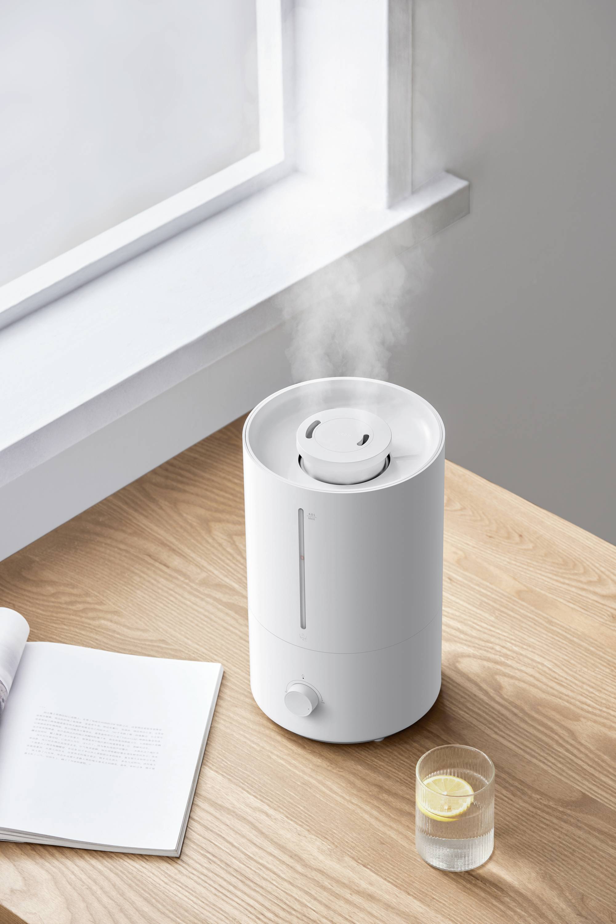 Un umidificatore bianco emana vapore su un tavolo di legno accanto a un libro aperto e un bicchiere d'acqua con una fetta di limone.