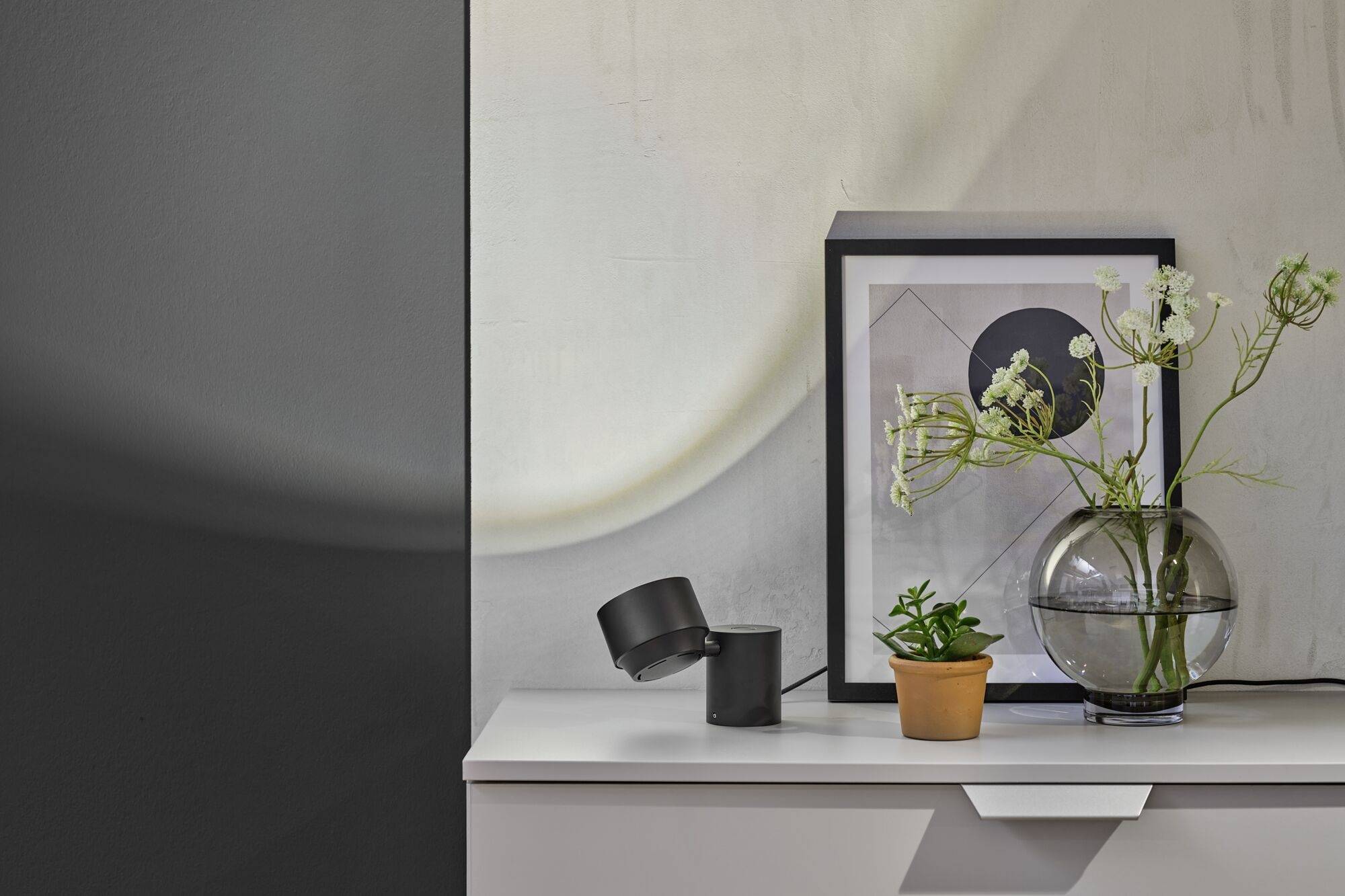 Una scaffalatura arredata in modo minimalista con un'immagine incorniciata, un vaso di vetro con fiori, un piccolo vaso da fiori e due cilindri neri.