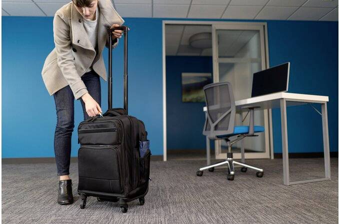 Una persona traina una valigia nera con rotelle in una stanza d'ufficio. Sullo sfondo, un tavolo con un laptop e una sedia da ufficio.