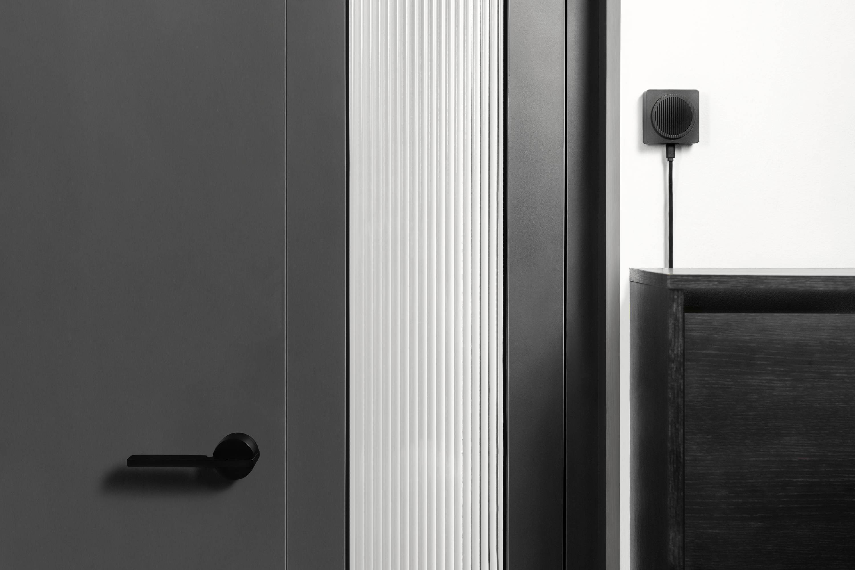 Una porta moderna e minimalista con maniglia nera, montata accanto a una parete bianca con un piccolo dispositivo sensore quadrato.