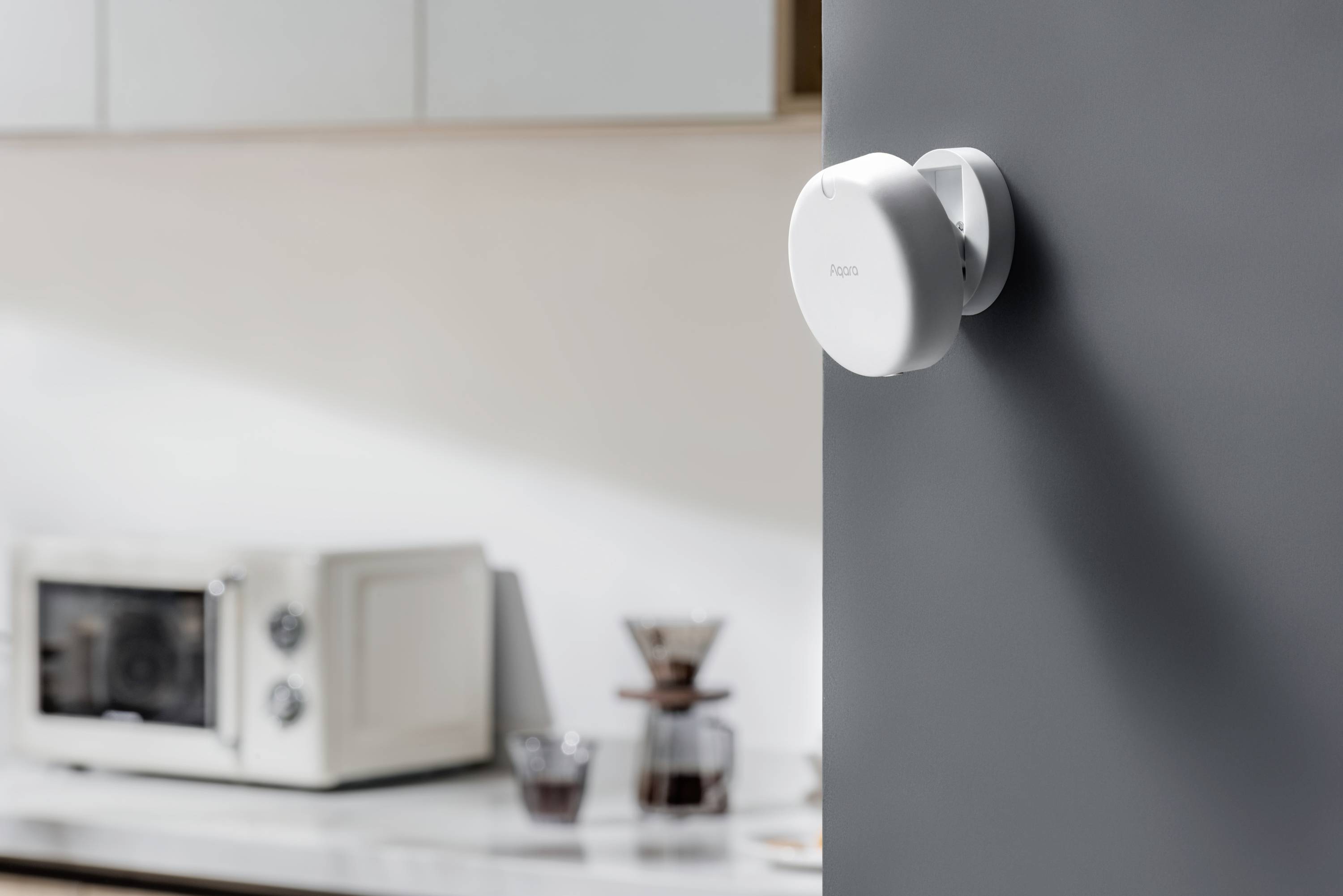 Un moderno termostato rotondo installato su una parete grigia in una cucina. Sullo sfondo sono visibili un forno a microonde e una macchina da caffè.