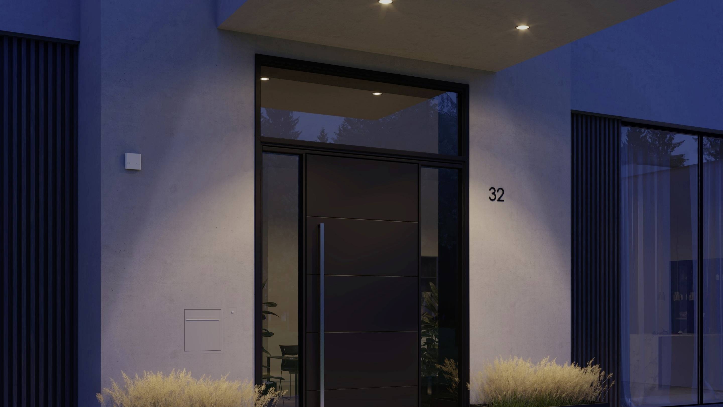 Moderna porta d'ingresso di una casa al crepuscolo, illuminata da tre faretti a soffitto. Numero civico '32' sulla parete accanto alla porta.