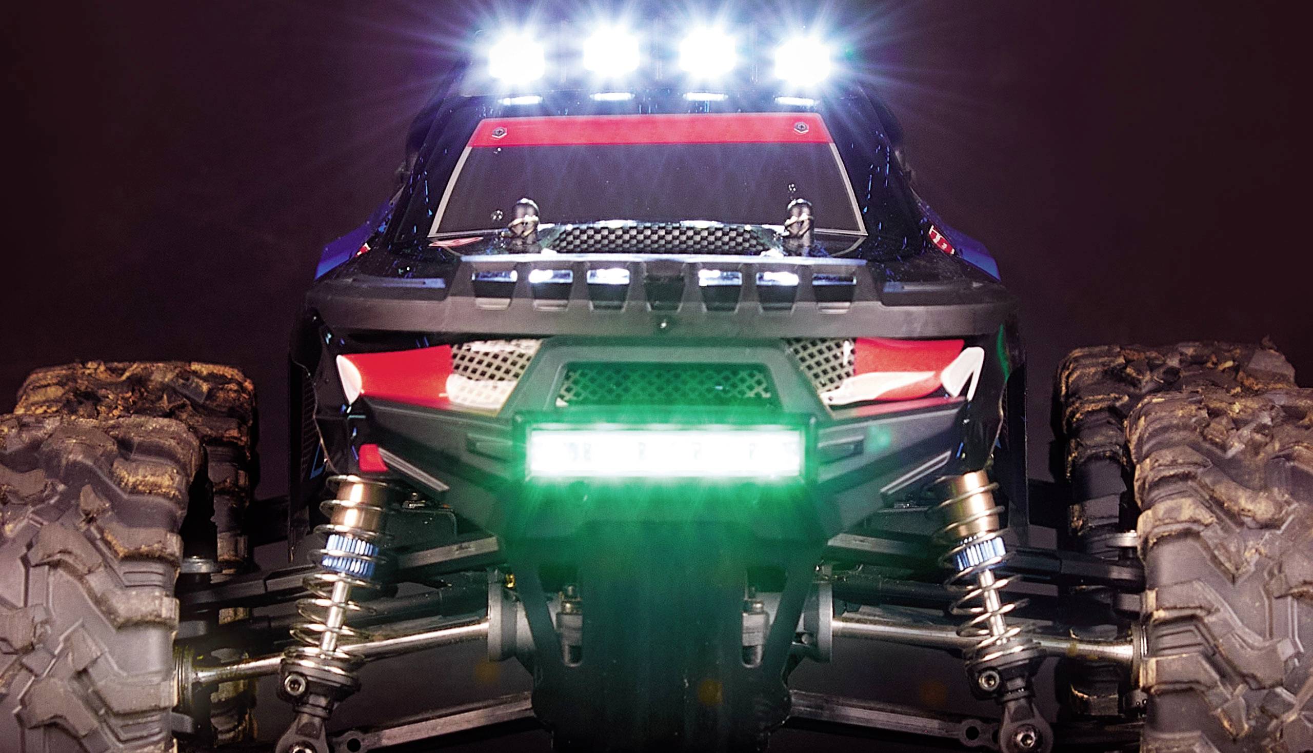Un'auto radiocomandata in primo piano, vista frontale, con luci LED illuminate, grandi pneumatici da fuoristrada e sospensioni visibili.