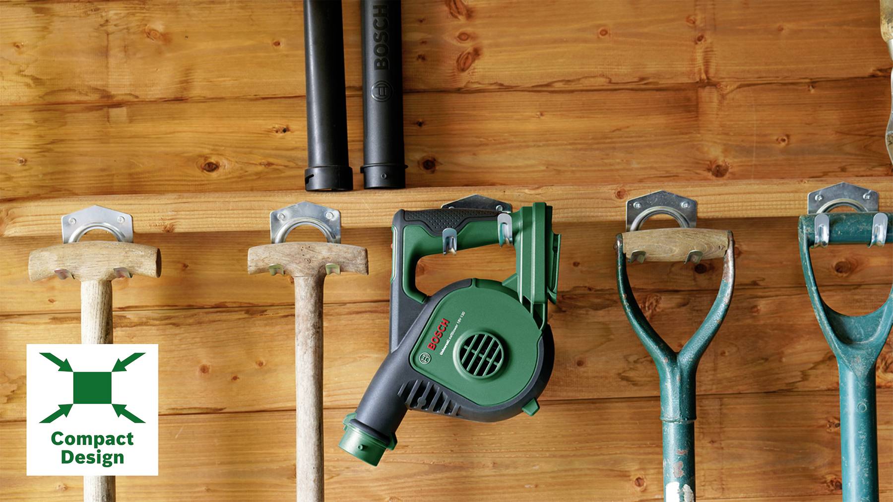 Un attrezzo da giardino verde con la scritta 'Compact Design' è appeso a una tavola di legno tra martelli e pale su una parete di legno.