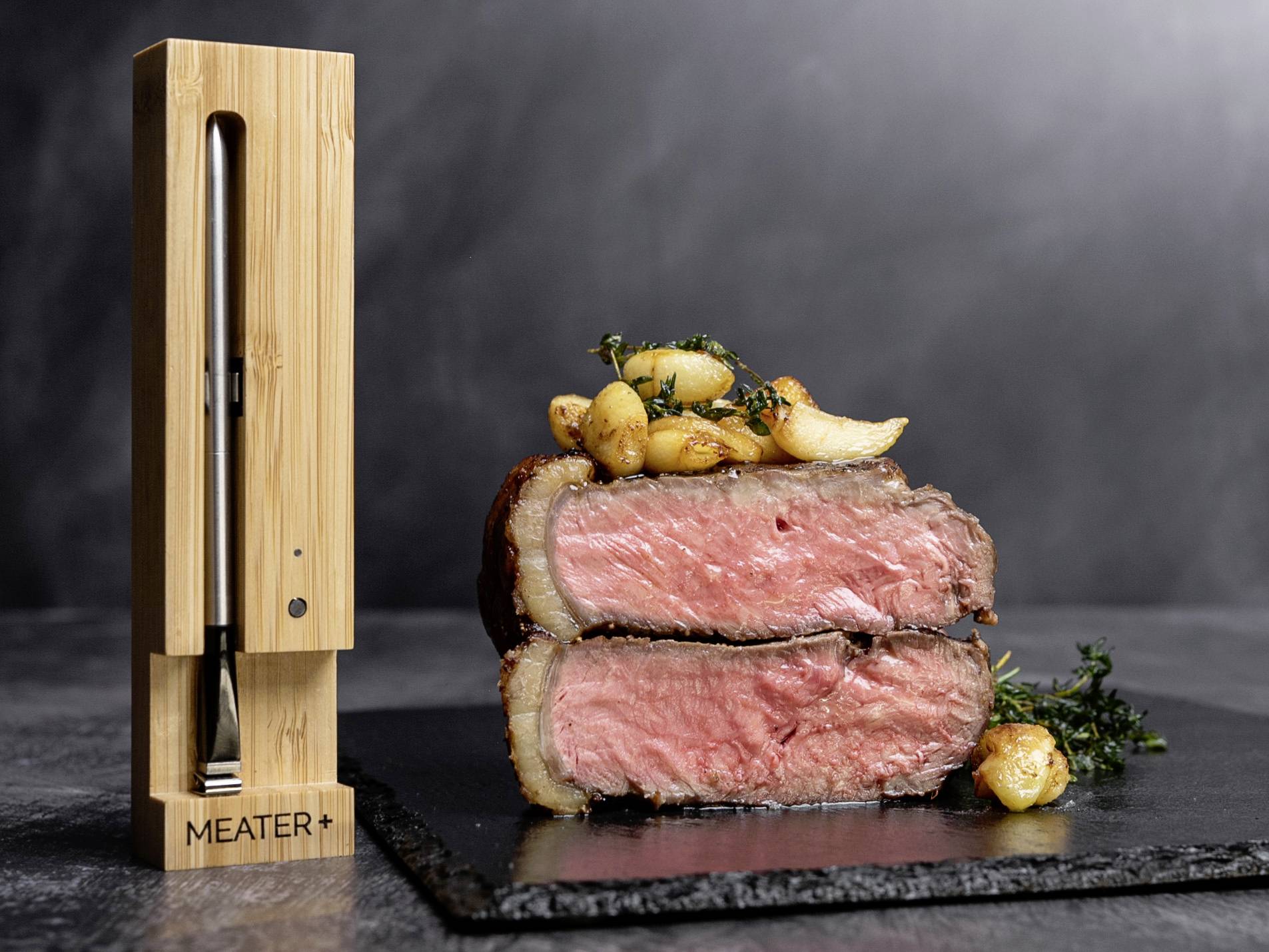 Un filetto di manzo tagliato e rosolato rosa su un piatto di ardesia, guarnito con erbe e aglio; accanto un termometro per la carne senza fili.