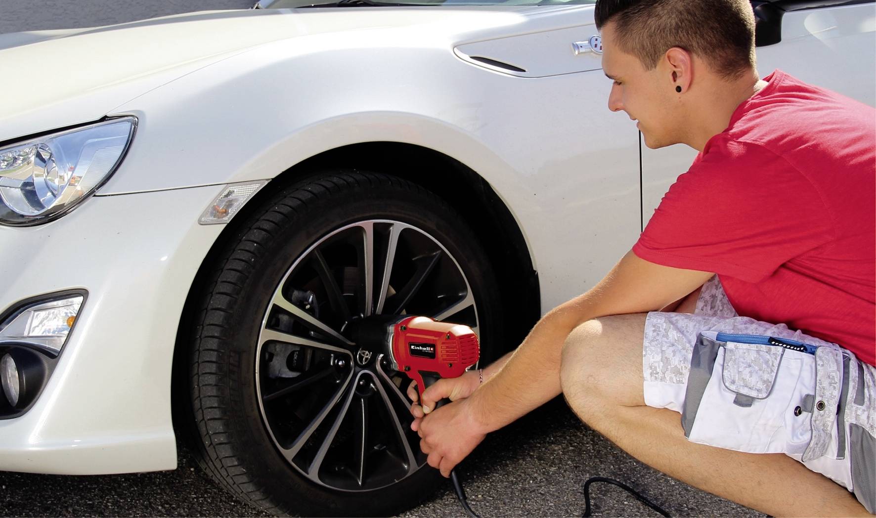 Un uomo con una maglietta rossa e pantaloncini corti sta cambiando un pneumatico di un'auto bianca usando una chiave inglese.