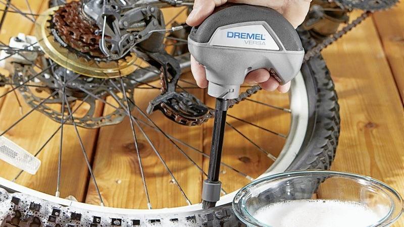Una persona sta pulendo una bicicletta con un dispositivo elettrico di pulizia chiamato 'Dremel Versa' accanto a una ciotola con acqua e sapone.