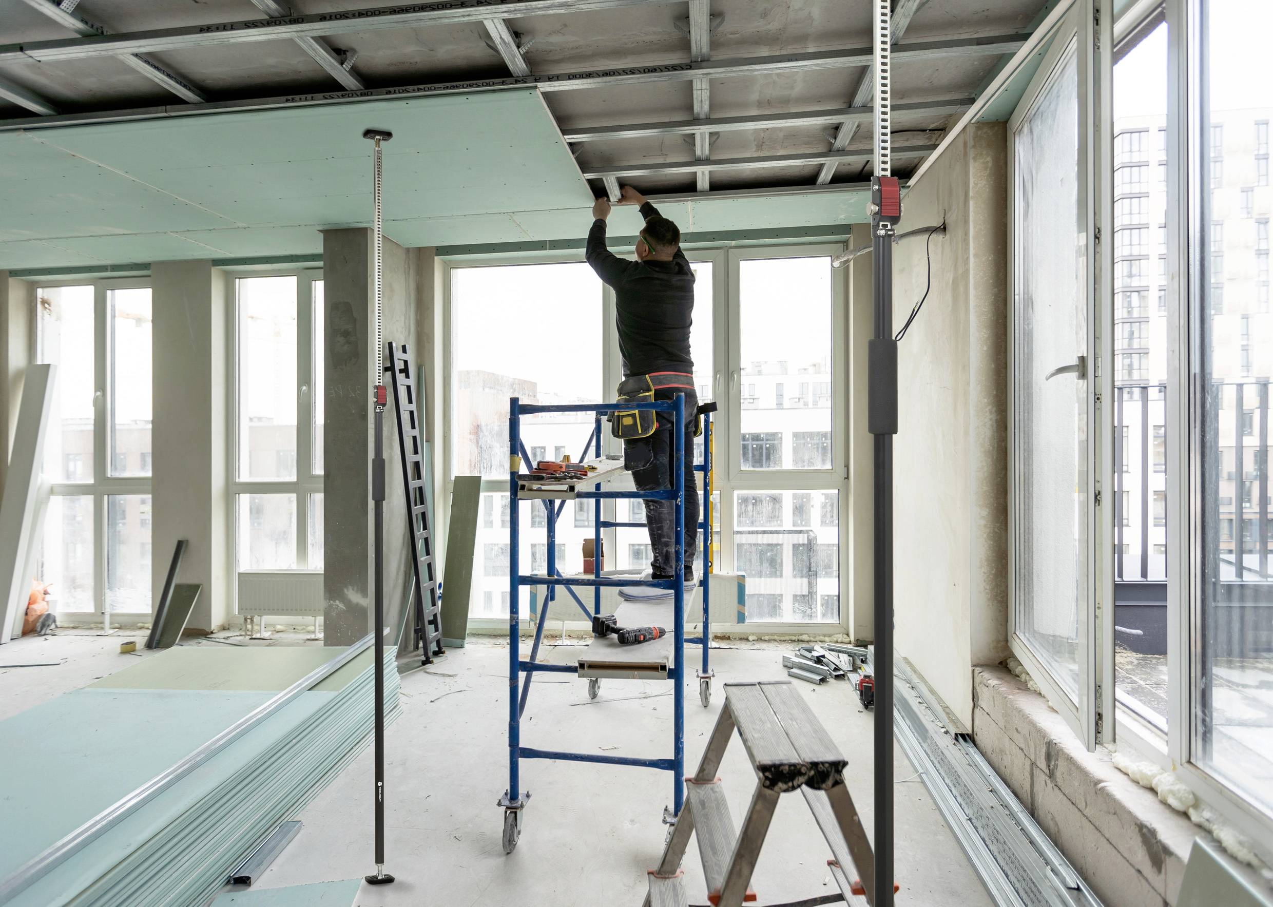 Una persona è in piedi su un ponteggio e sta montando lastre di cartongesso sul soffitto di una stanza non finita con grandi finestre.