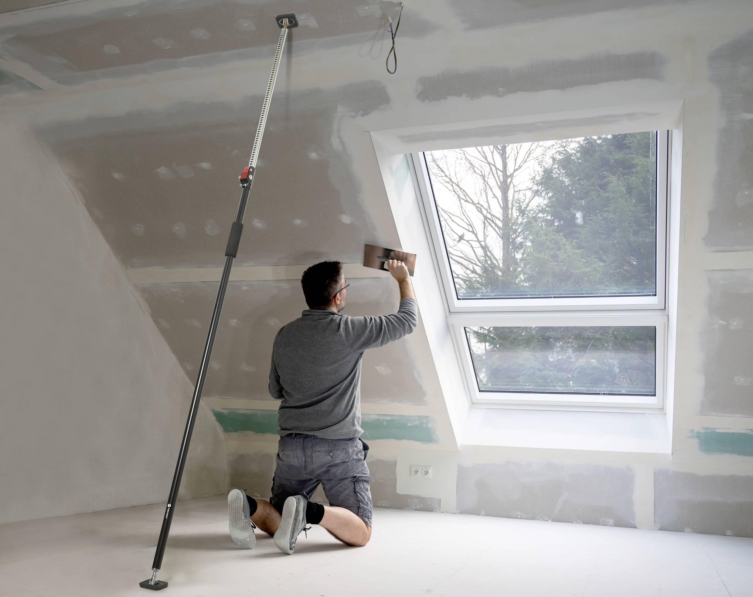 Un uomo è inginocchiato e stucca un tetto inclinato. Accanto a lui un finestra con vista su degli alberi. Un'asta telescopica sorregge il soffitto.