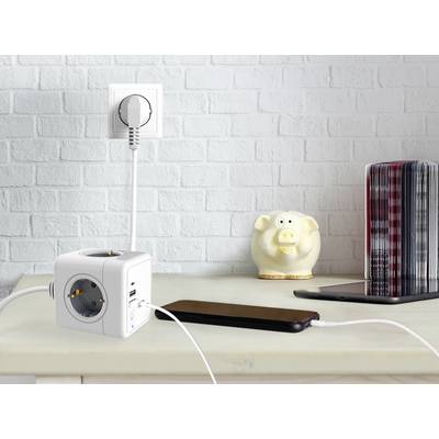 Un adattatore multipresa a cubo con due porte USB sta caricando uno smartphone. Accanto, un salvadanaio è poggiato su uno scaffale.