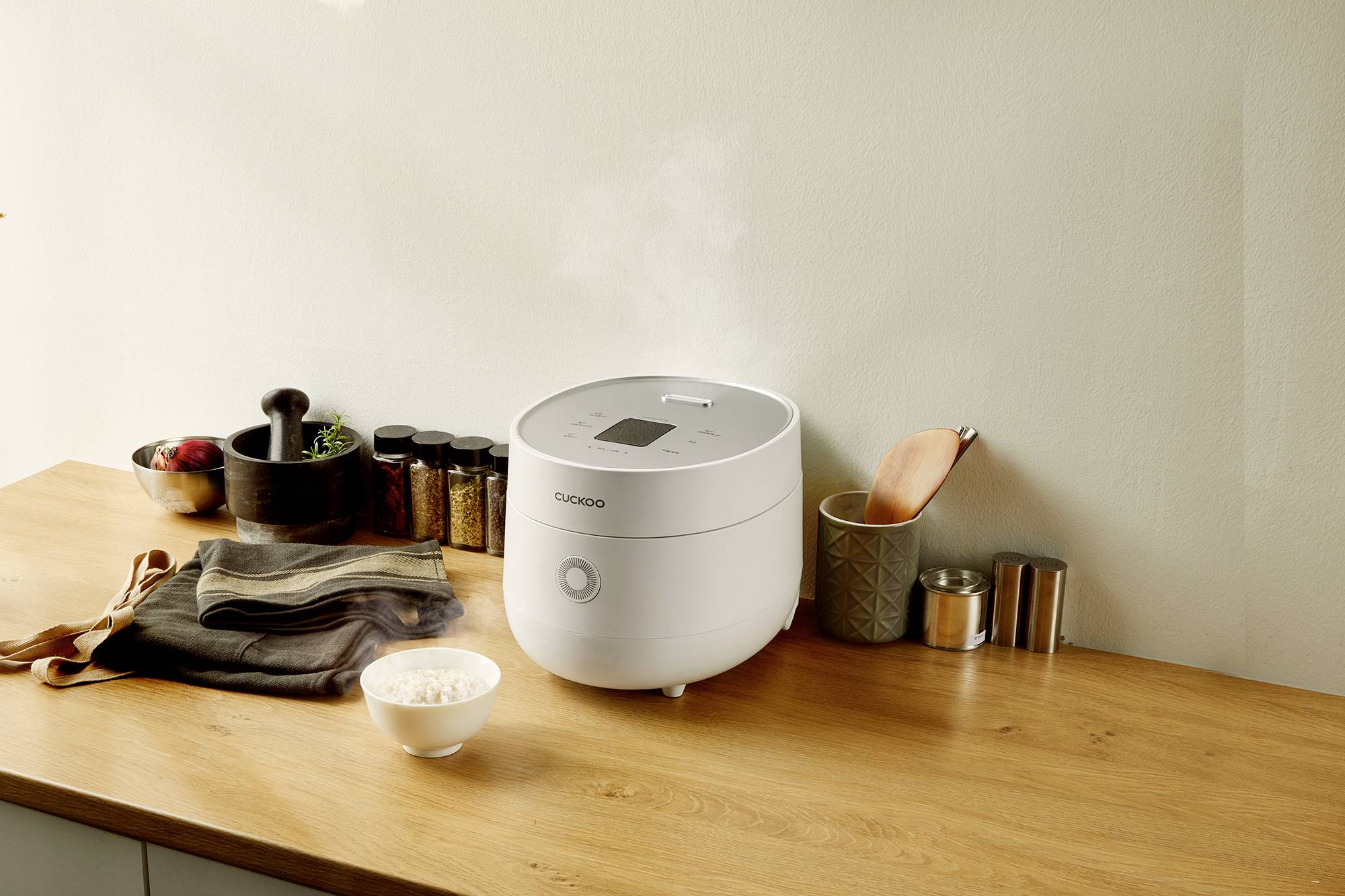 Un fornello di riso bianco sta emettendo vapore su un bancone di legno. Accanto a esso una ciotola di riso, utensili da cucina e vasetti di spezie. Ambiente di cucina.