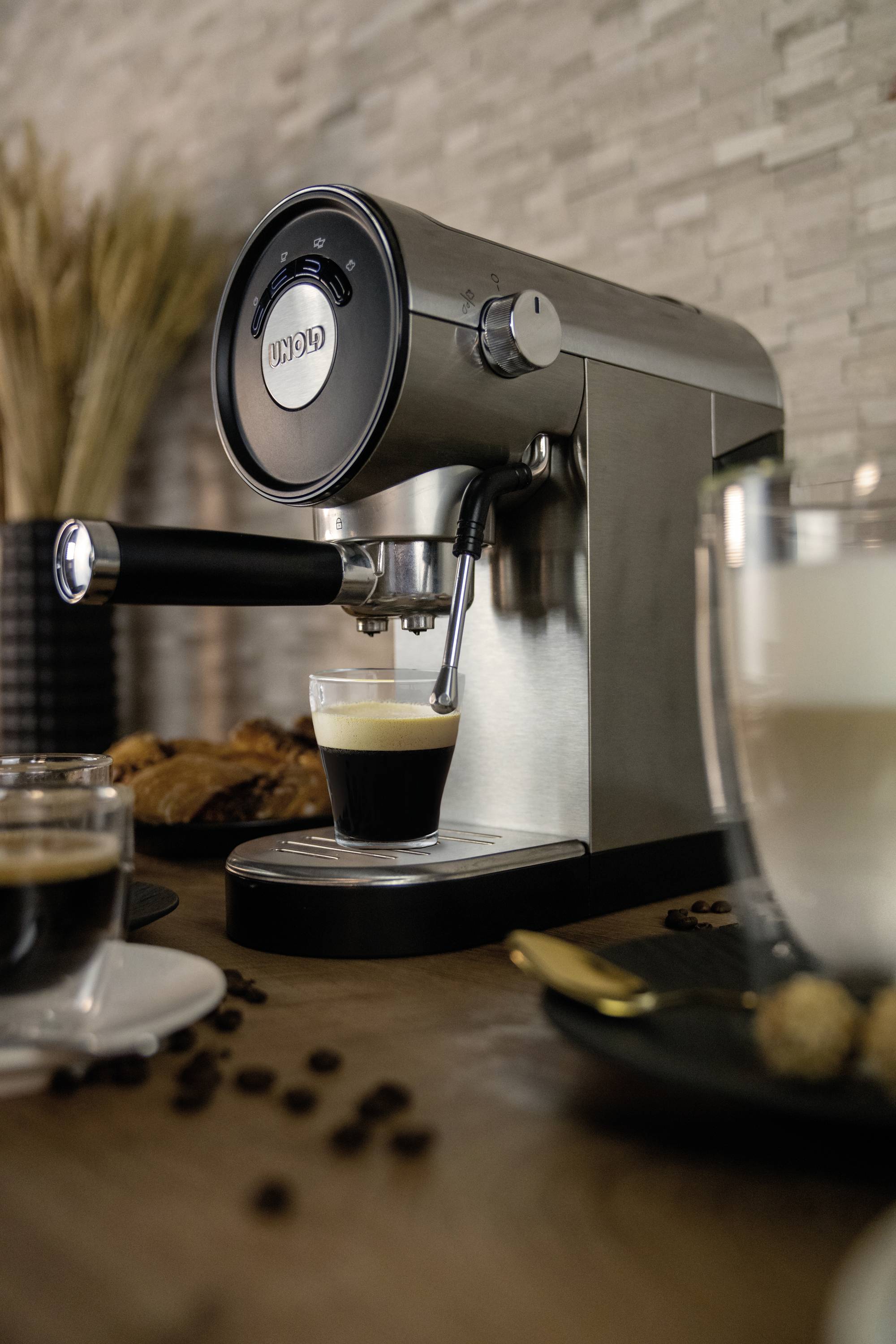 Una macchina per espresso argentata su un piano di lavoro, con un piccolo bicchiere di espresso sotto di essa. Sullo sfondo sono sparse alcune chicche di caffè.