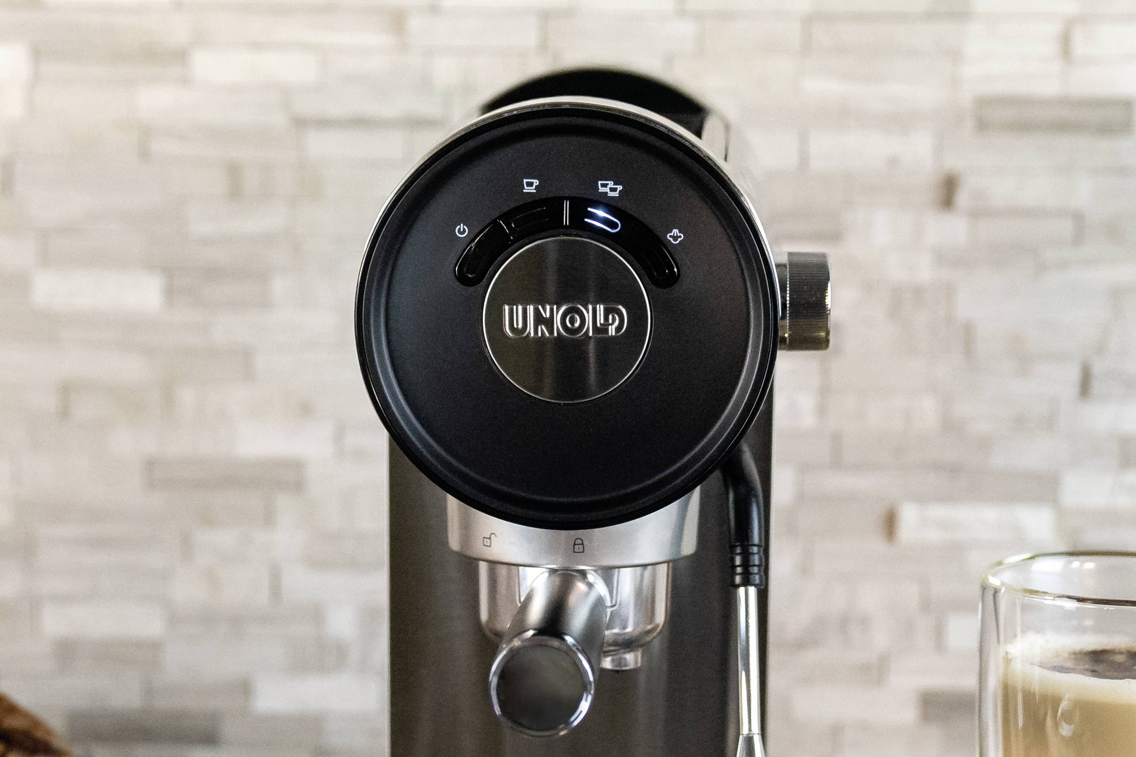 Una macchina per espresso in argento con pannello di controllo nero e la scritta 'UNOLD', davanti a un muro piastrellato. A destra, un bicchiere di latte.