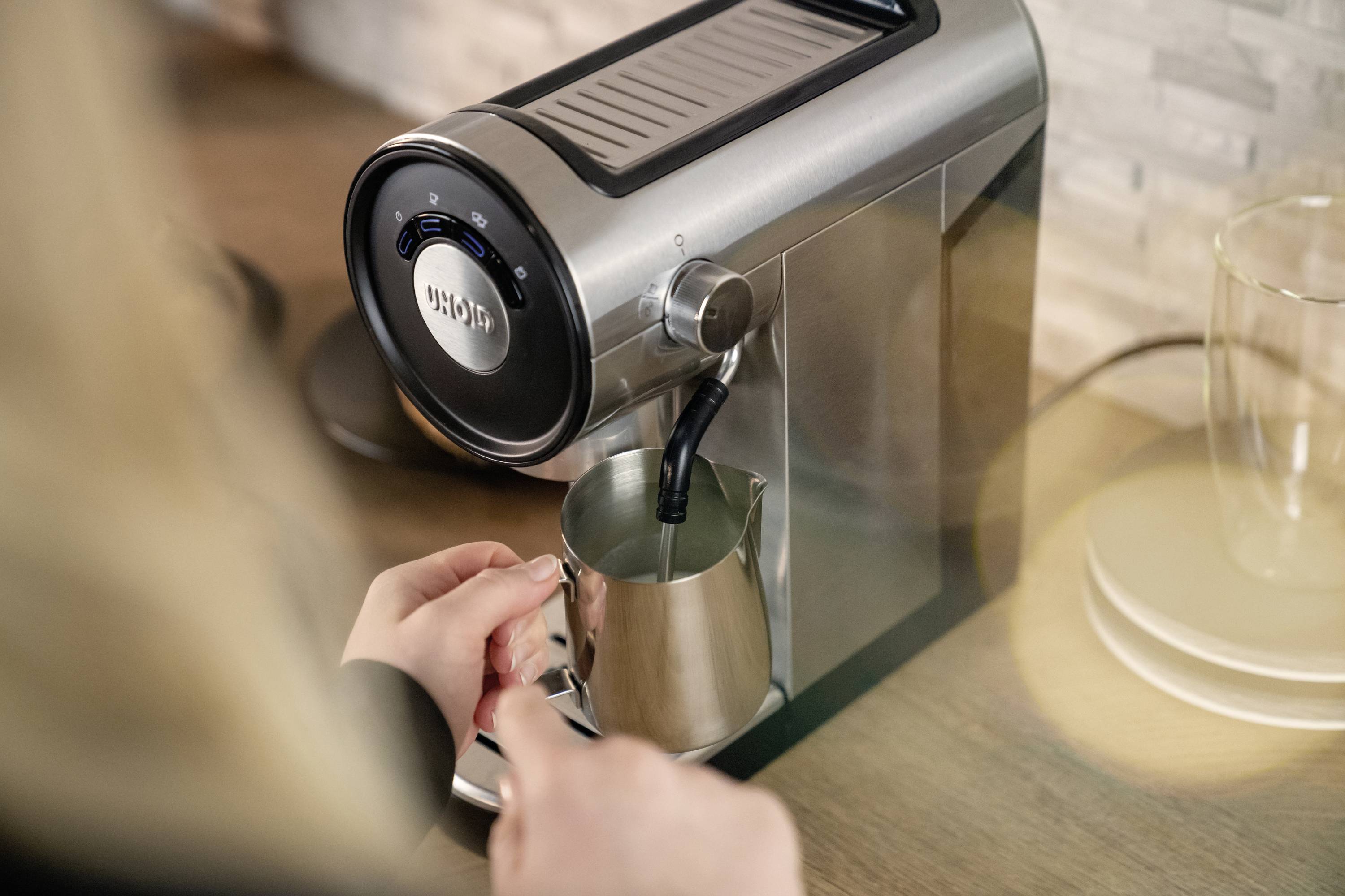 Una persona sta montando il latte con una macchina da caffè argentata in una brocca di acciaio inossidabile. Sullo sfondo c'è un piano di lavoro da cucina.