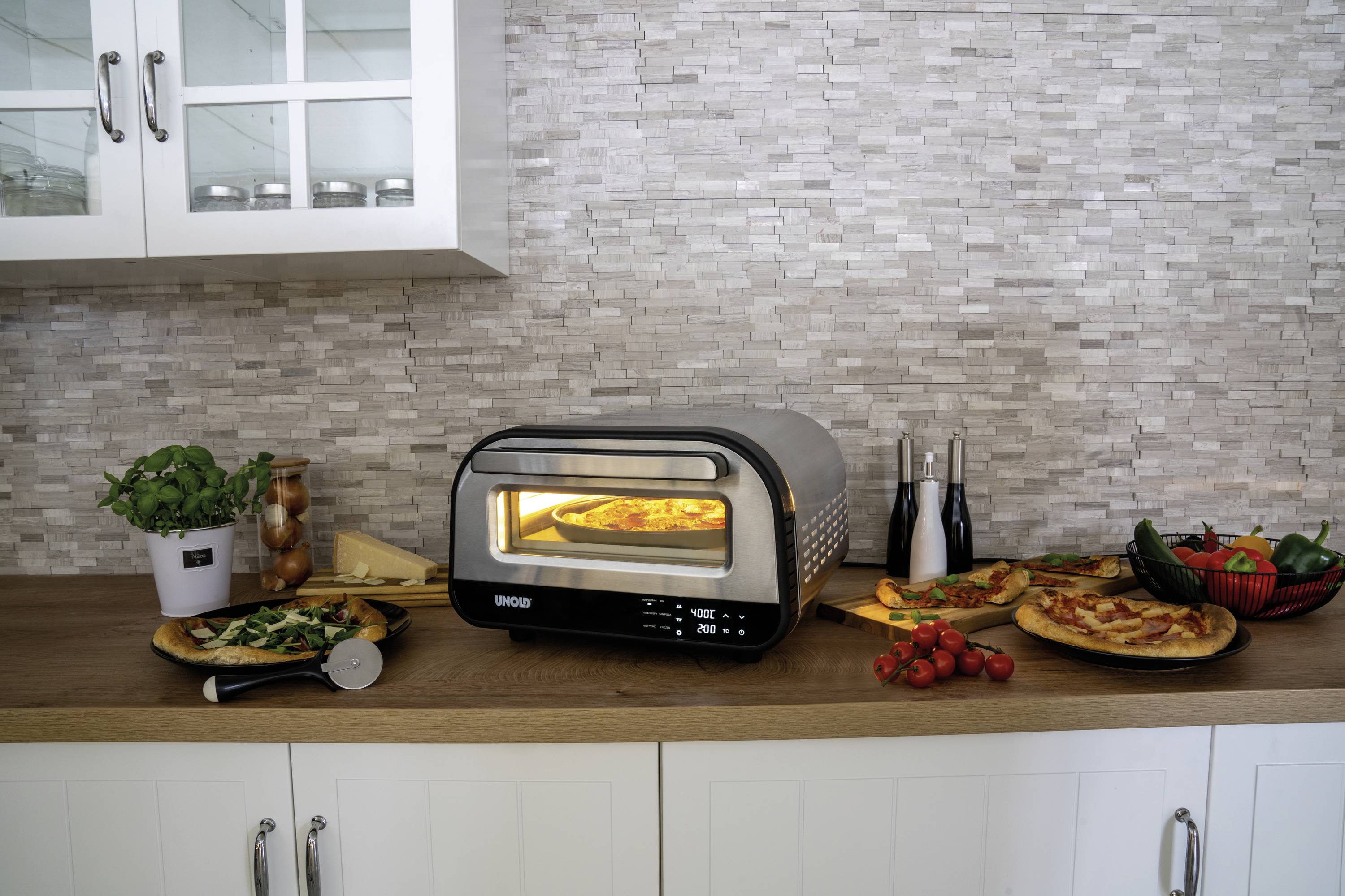 Un forno moderno su un piano di lavoro della cucina cuoce una pizza. Intorno ad esso ci sono ingredienti freschi e fette di pizza già pronte su piatti.