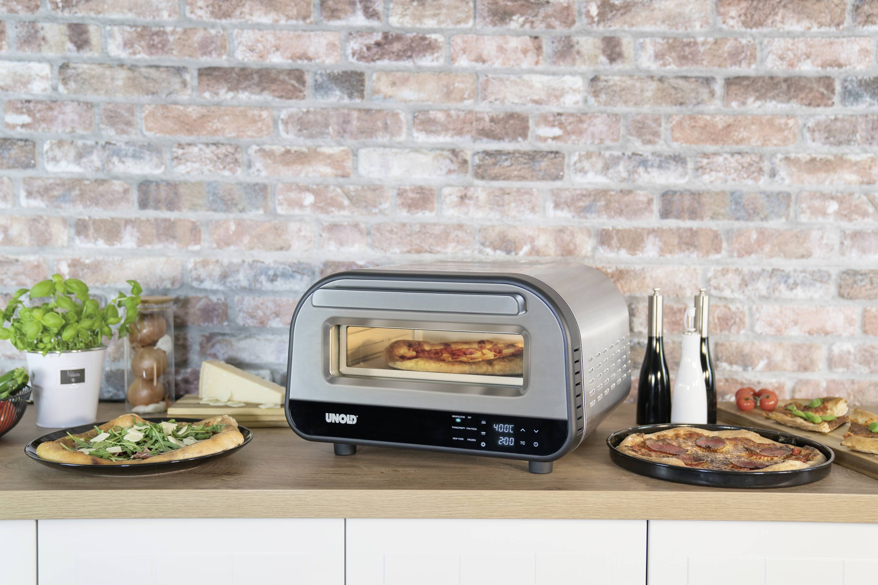 Un forno compatto su un piano di lavoro sta cuocendo una pizza. Accanto ci sono pizze già cotte. Sullo sfondo, un muro di mattoni.